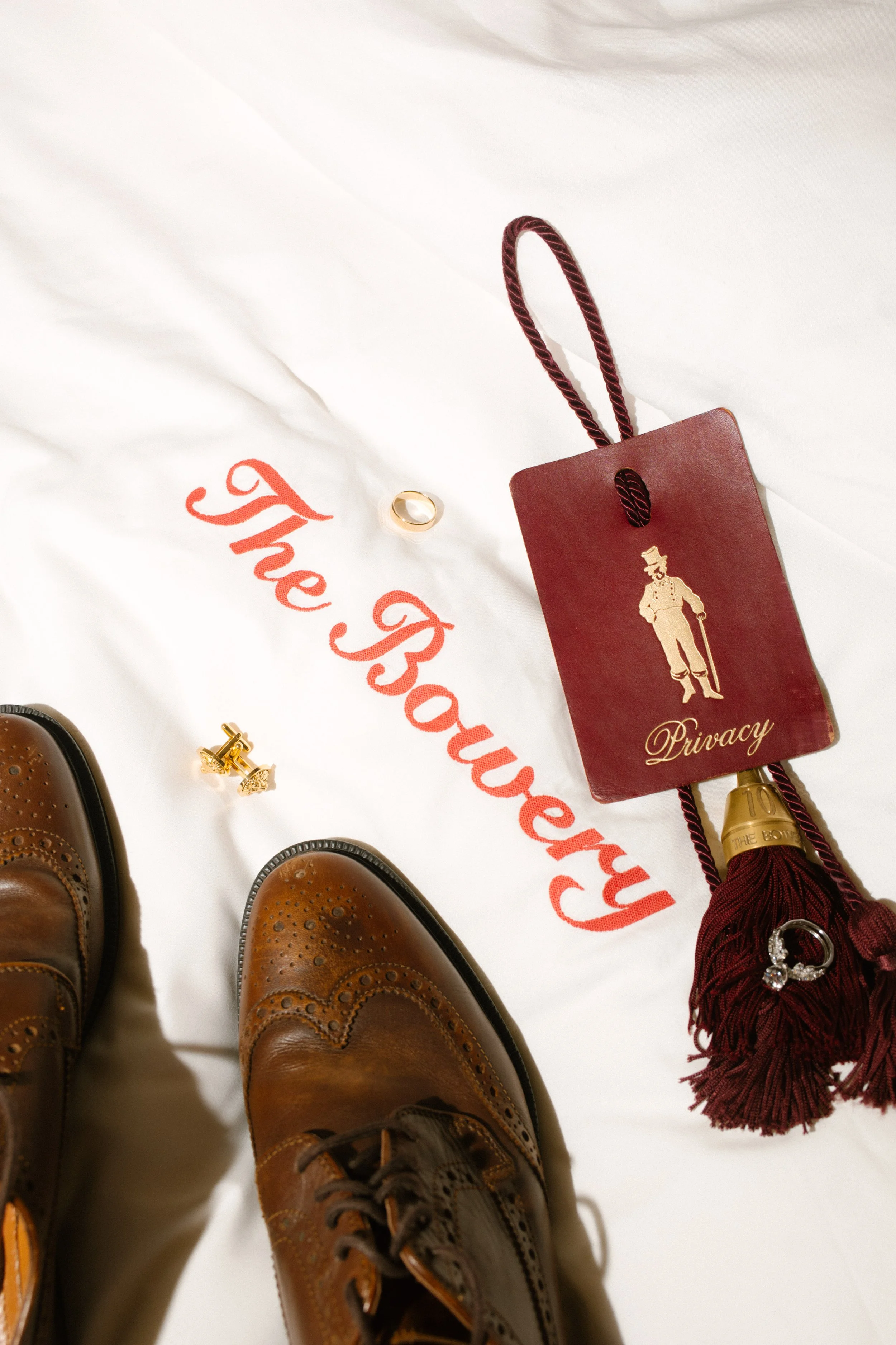 Wedding-day flat lay featuring brown dress shoes, rings, cufflinks and The Bowery Hotel details.
