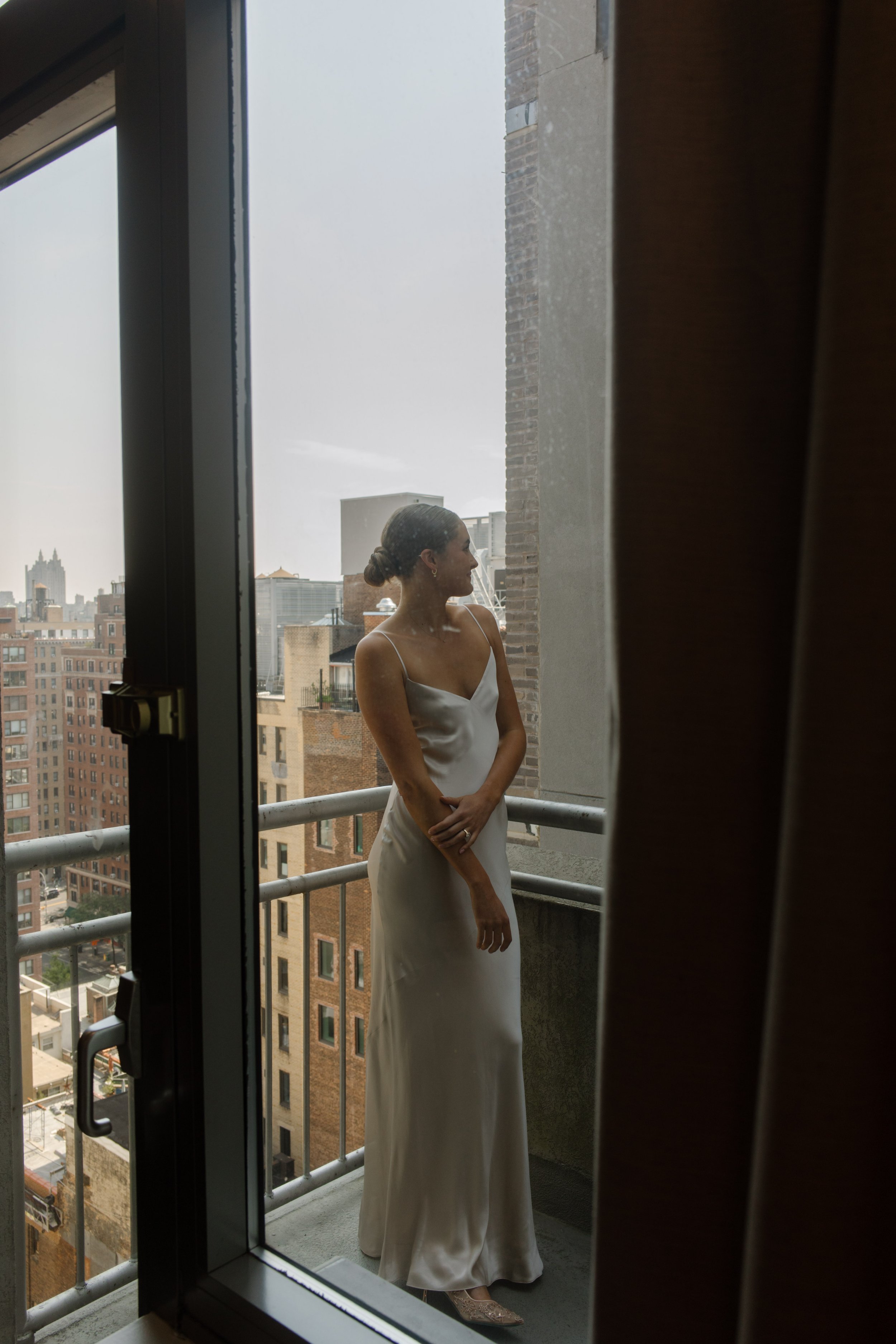 Bride in a satin wedding dress posing on a hotel balcony overlooking the New York City skyline.