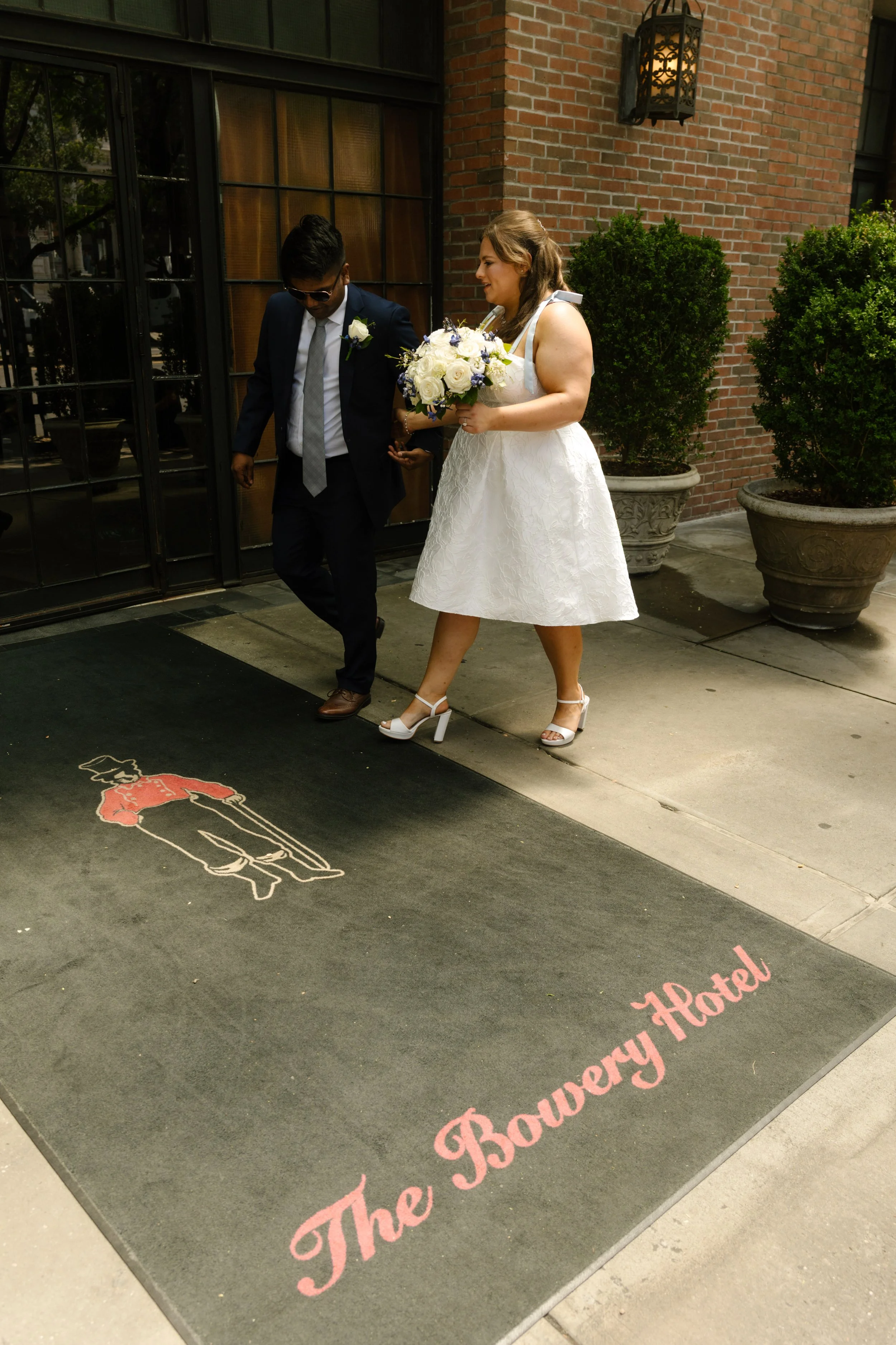 Newlyweds leaving The Bowery Hotel together, with the bride carrying a white-and-blue bouquet.