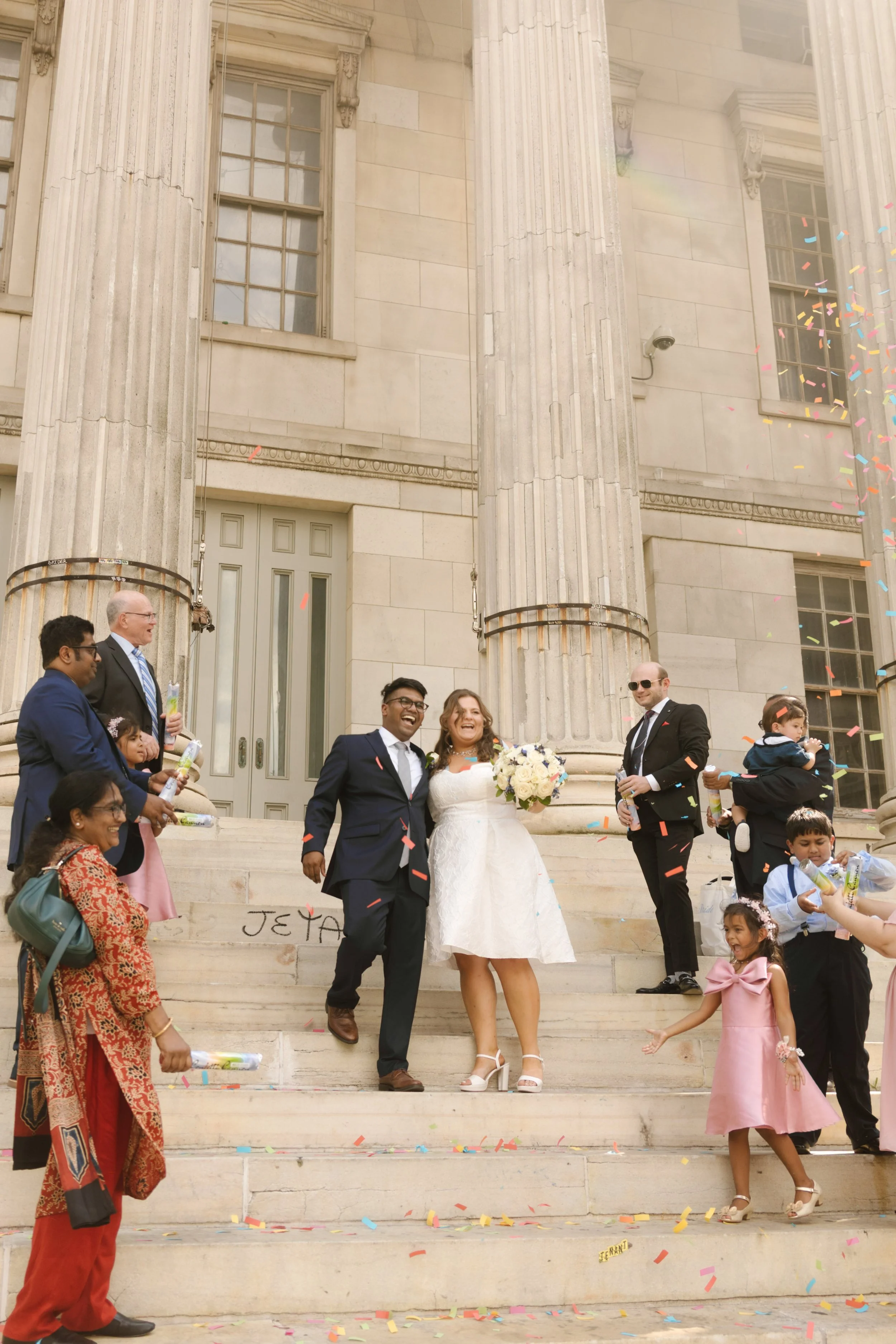 Newlyweds celebrating with family and friends under colorful confetti on courthouse steps in NYC.