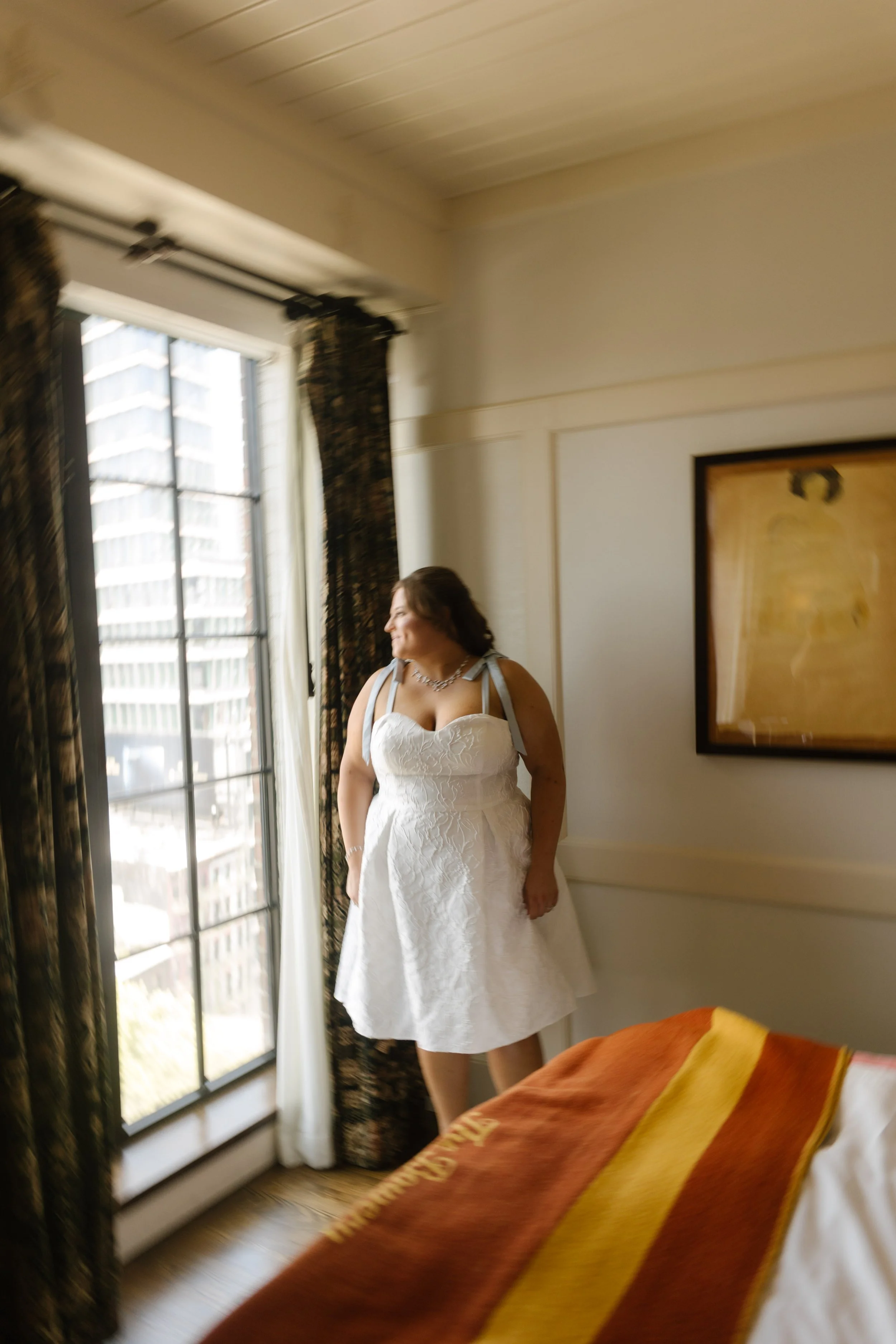 Bride in a white wedding dress looking out a large hotel room window.