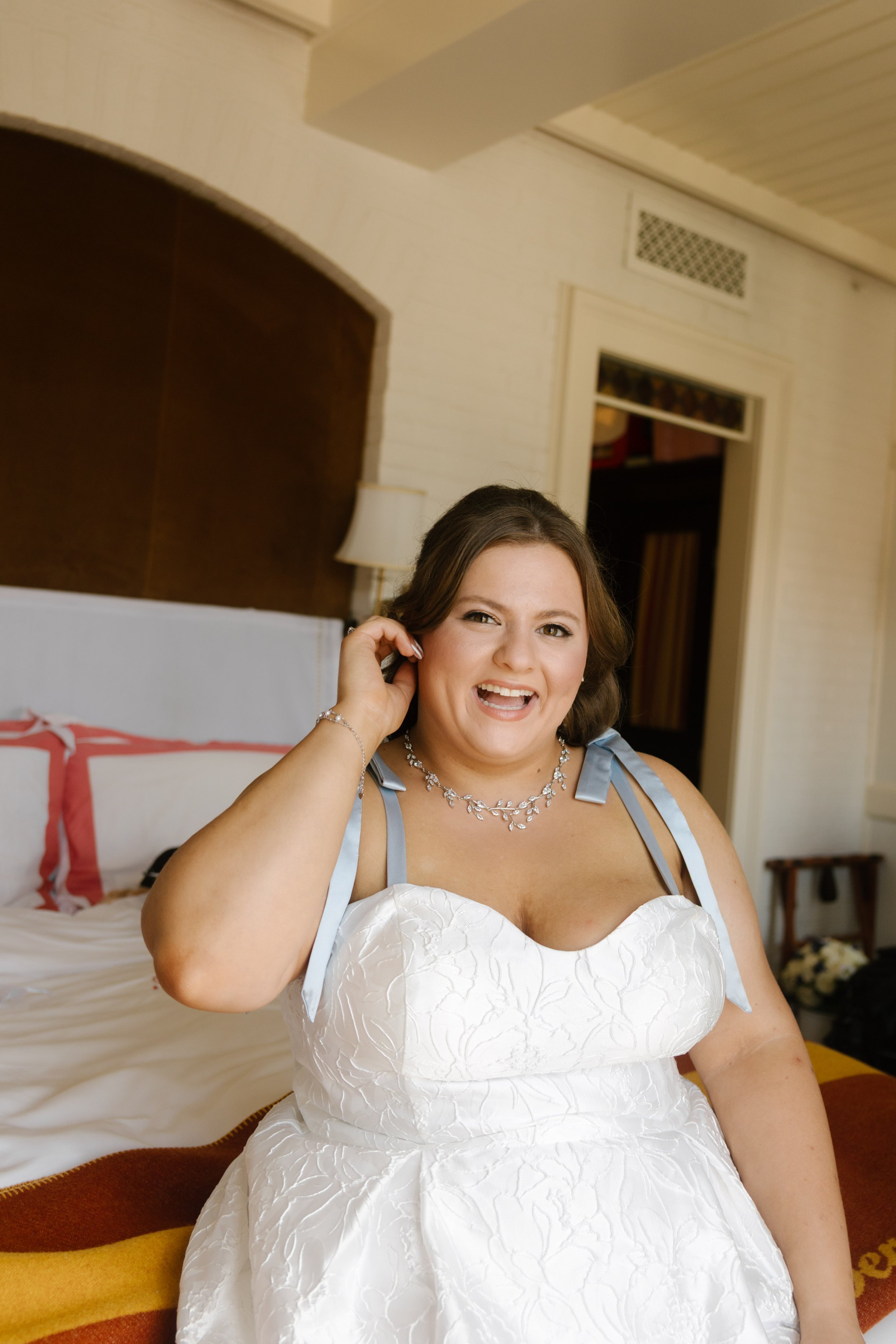 Smiling bride adjusting her earring while getting ready in a hotel room.