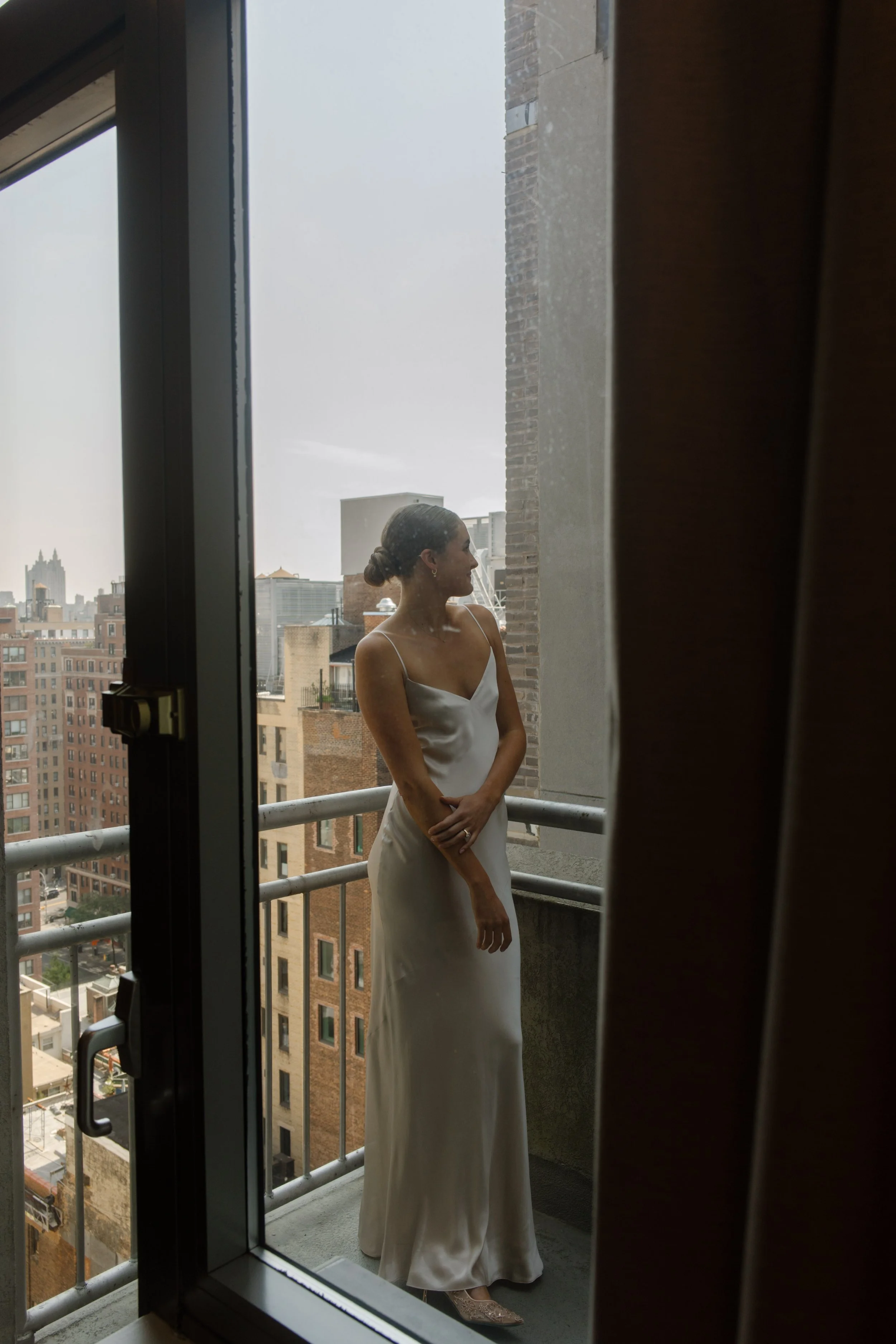 Bride in a satin wedding dress standing on a balcony overlooking the city from one of the NYC hotels.