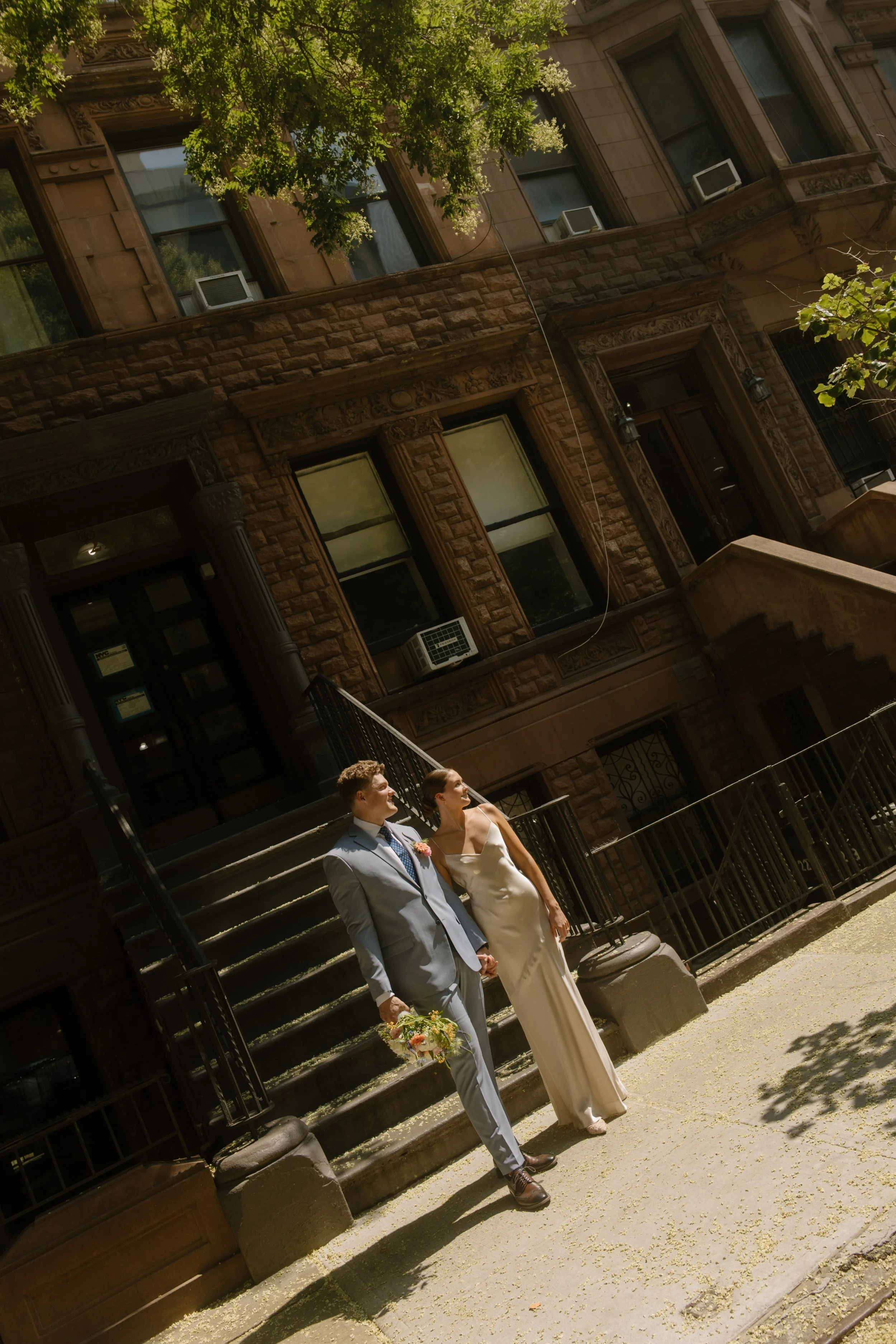 Newlyweds holding hands and posing on a sunlit New York City sidewalk in front of a brownstone.