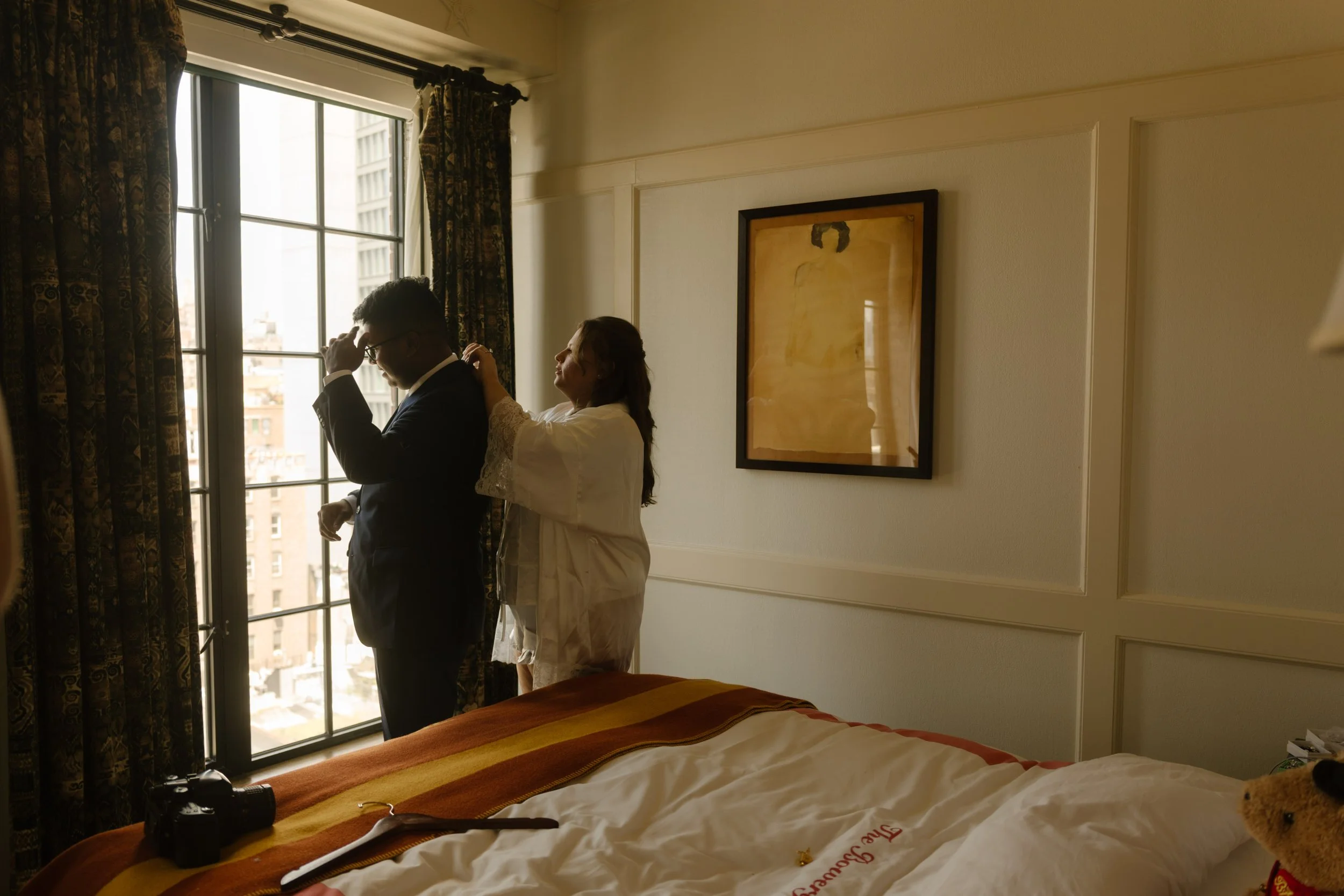 Bride helping the groom get ready beside a large window in a warmly decorated hotel room.