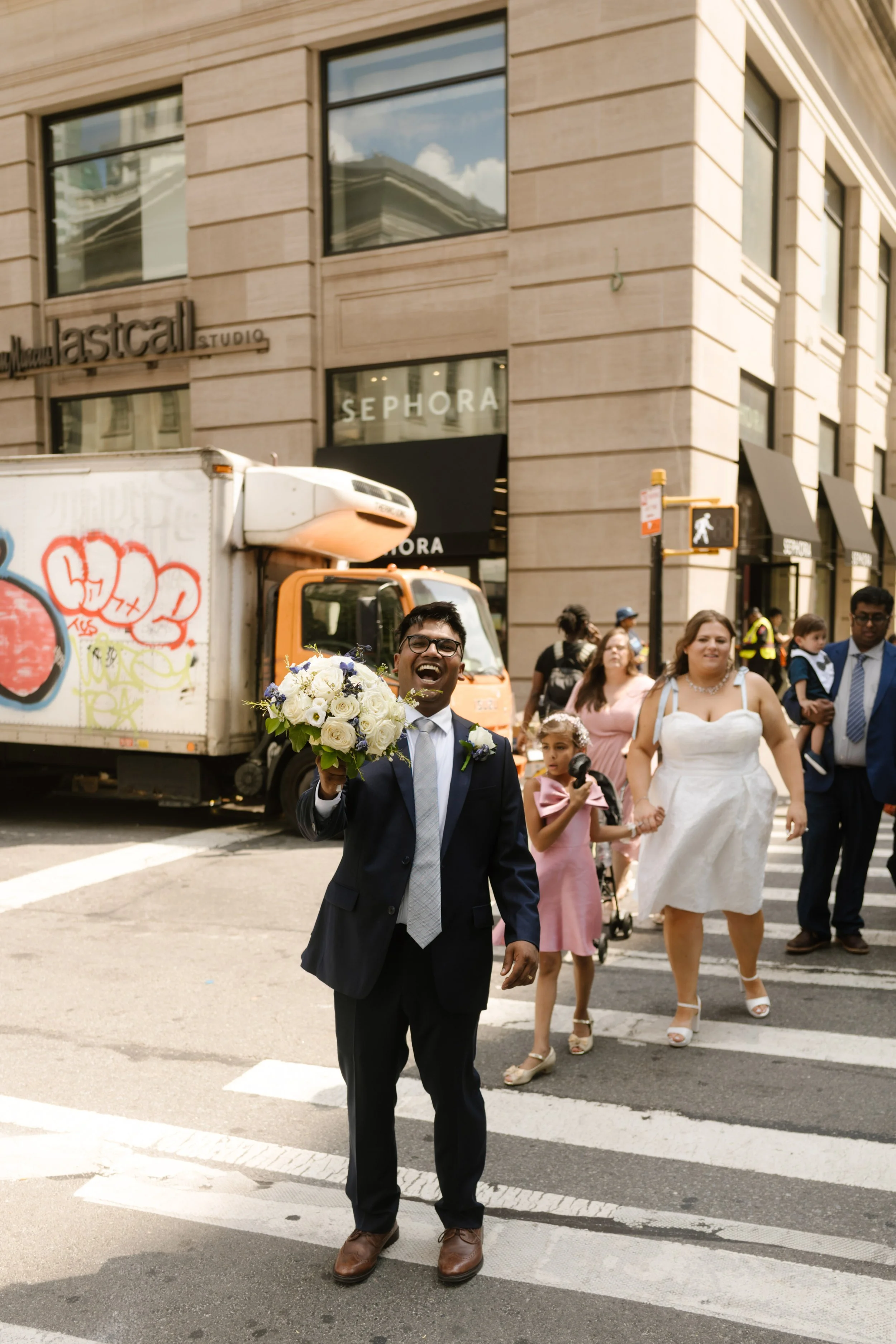 Groom carries his bouquet while crossing a busy Manhattan street with family after a New York City Hall wedding.