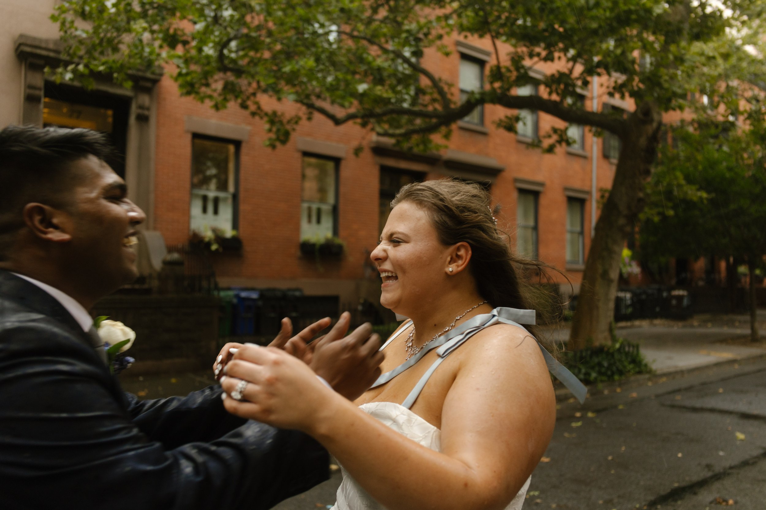 Bride and groom laughing together during rainy wedding portraits on a brownstone-lined New York street.