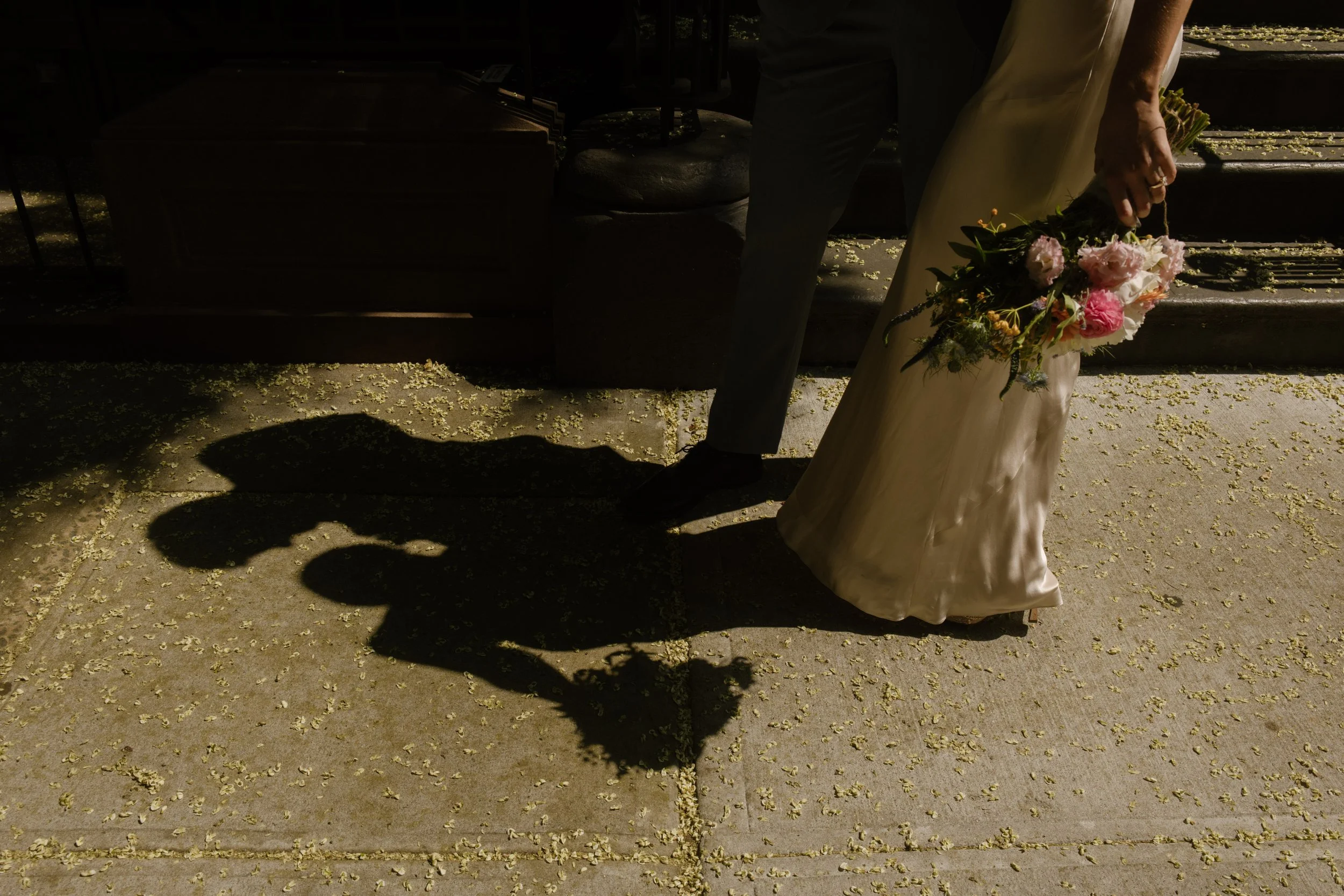Bride and groom walking in sunlight with a floral bouquet, casting dramatic shadows outside NYC hotels.