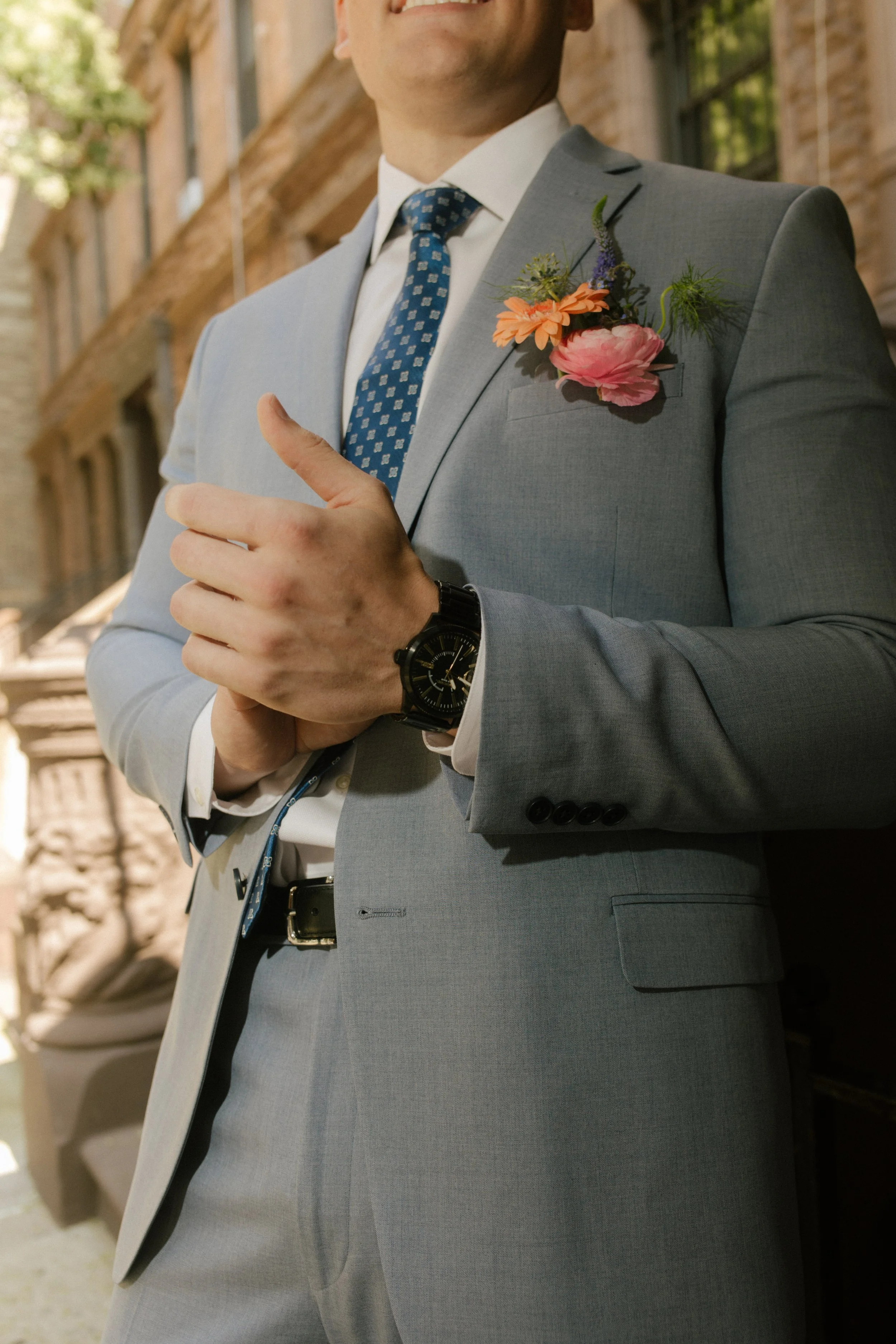 Close-up of a groom in a light gray suit with a colorful floral boutonniere and blue patterned tie.