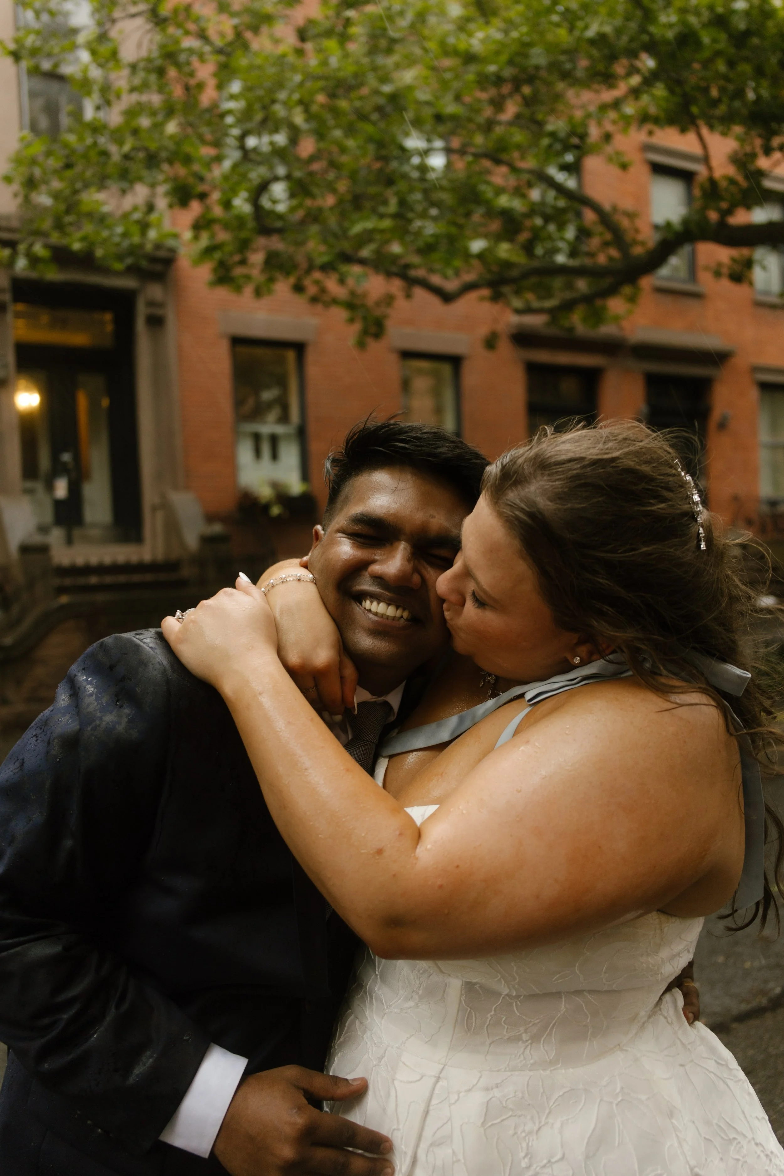 Bride kisses groom on the cheek during rainy wedding portraits on a tree-lined New York street.