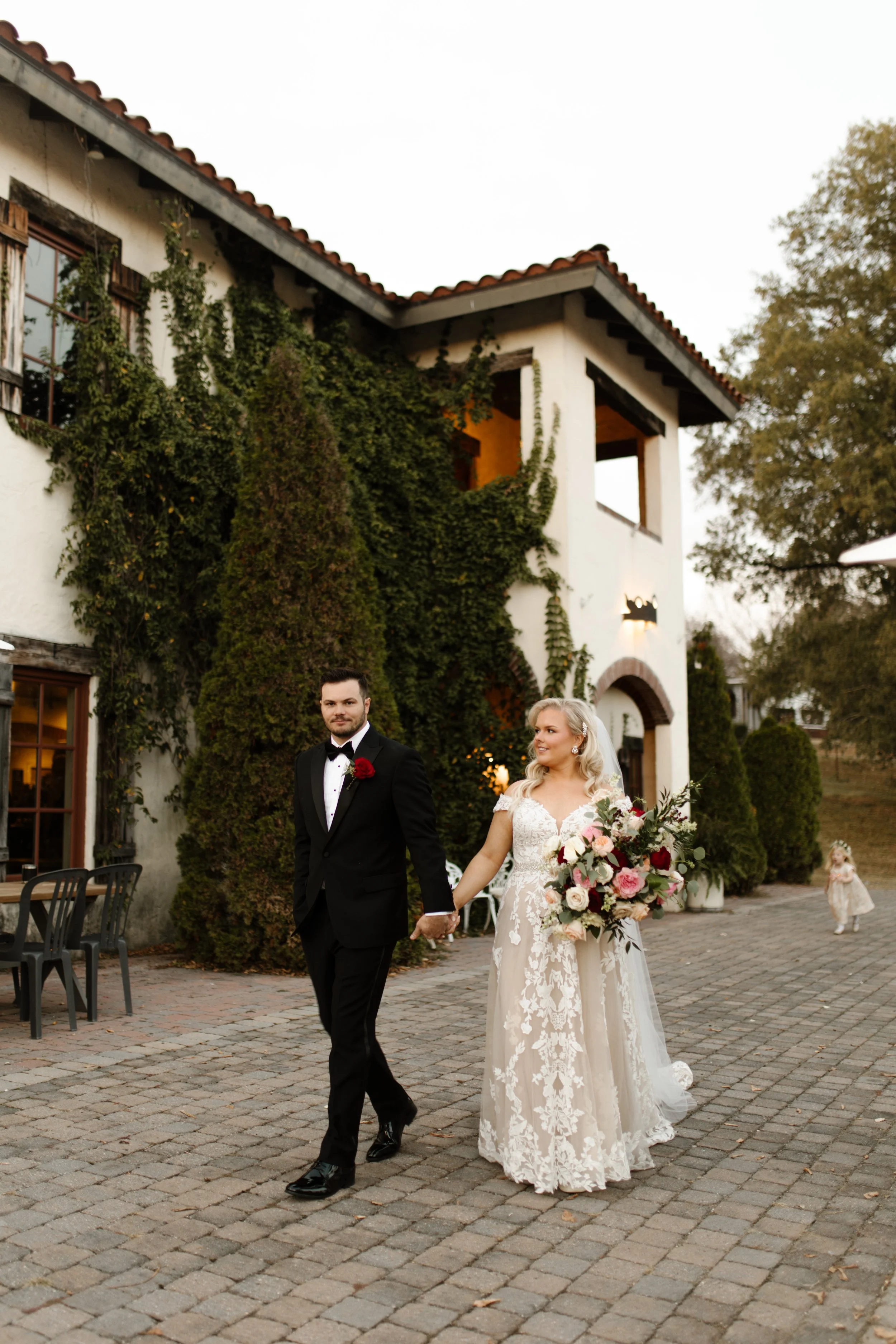 Bride and groom walking hand in hand outside an ivy-covered wedding venue with their flower girl behind them