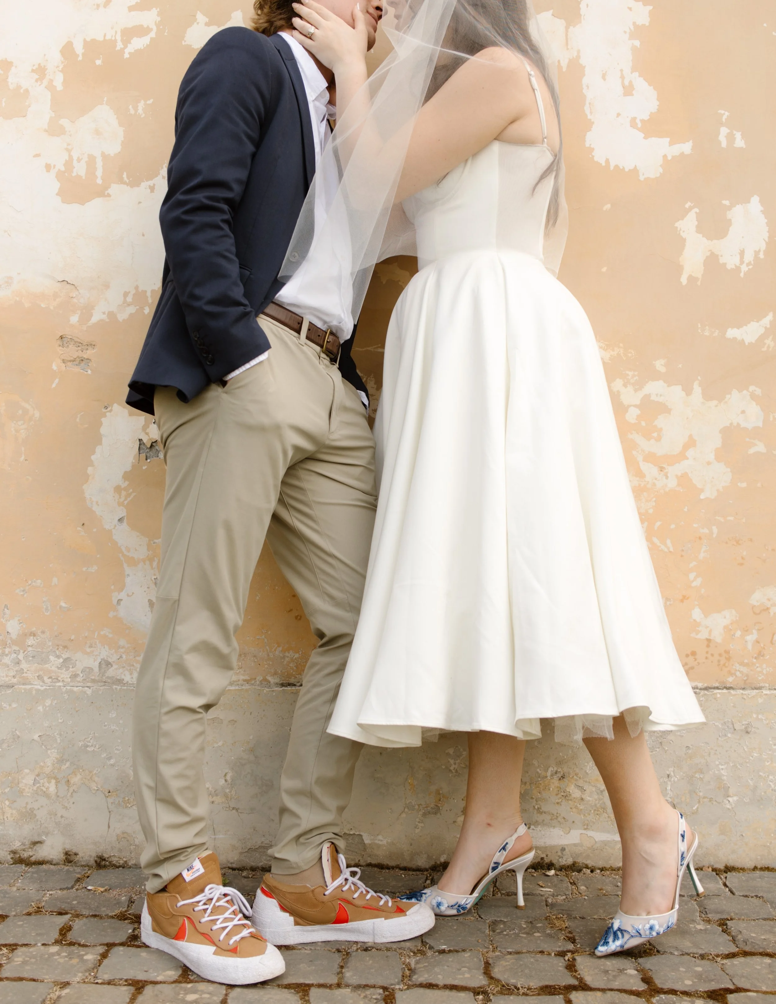 Newlywed couple kissing against a warm textured villa wall with the bride wearing colorful heels