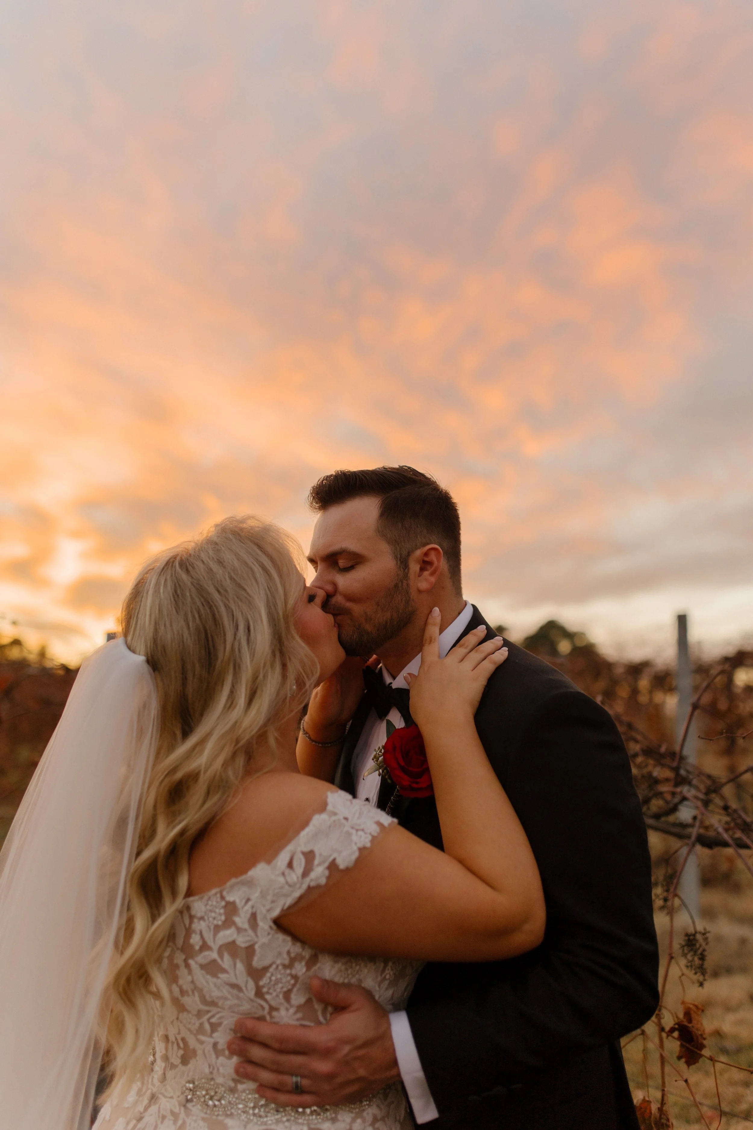 Bride and groom kissing in a vineyard beneath a colorful sunset during their destination wedding portraits