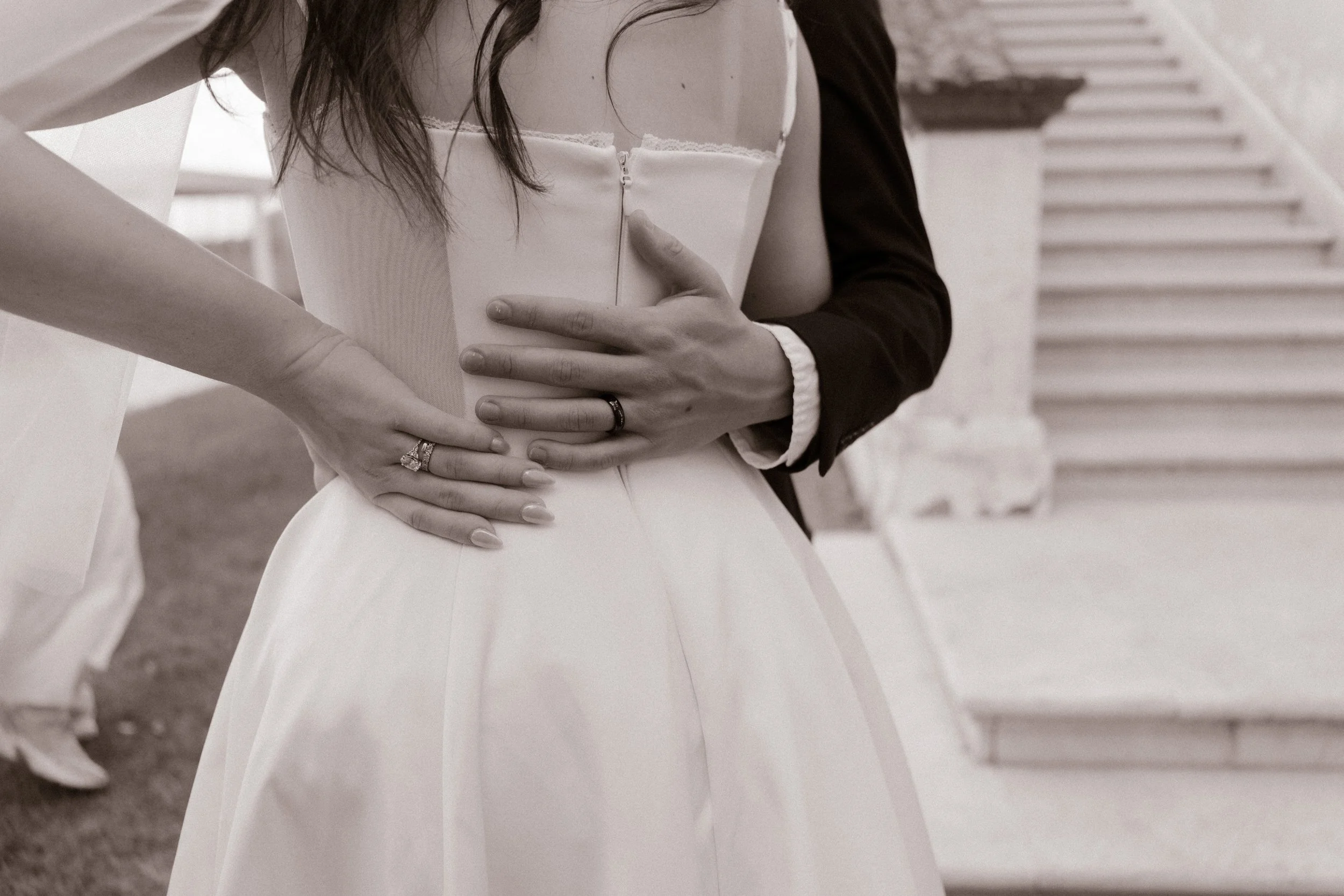 Black-and-white close-up of a newlywed couple embracing, showing their wedding rings and bridal details