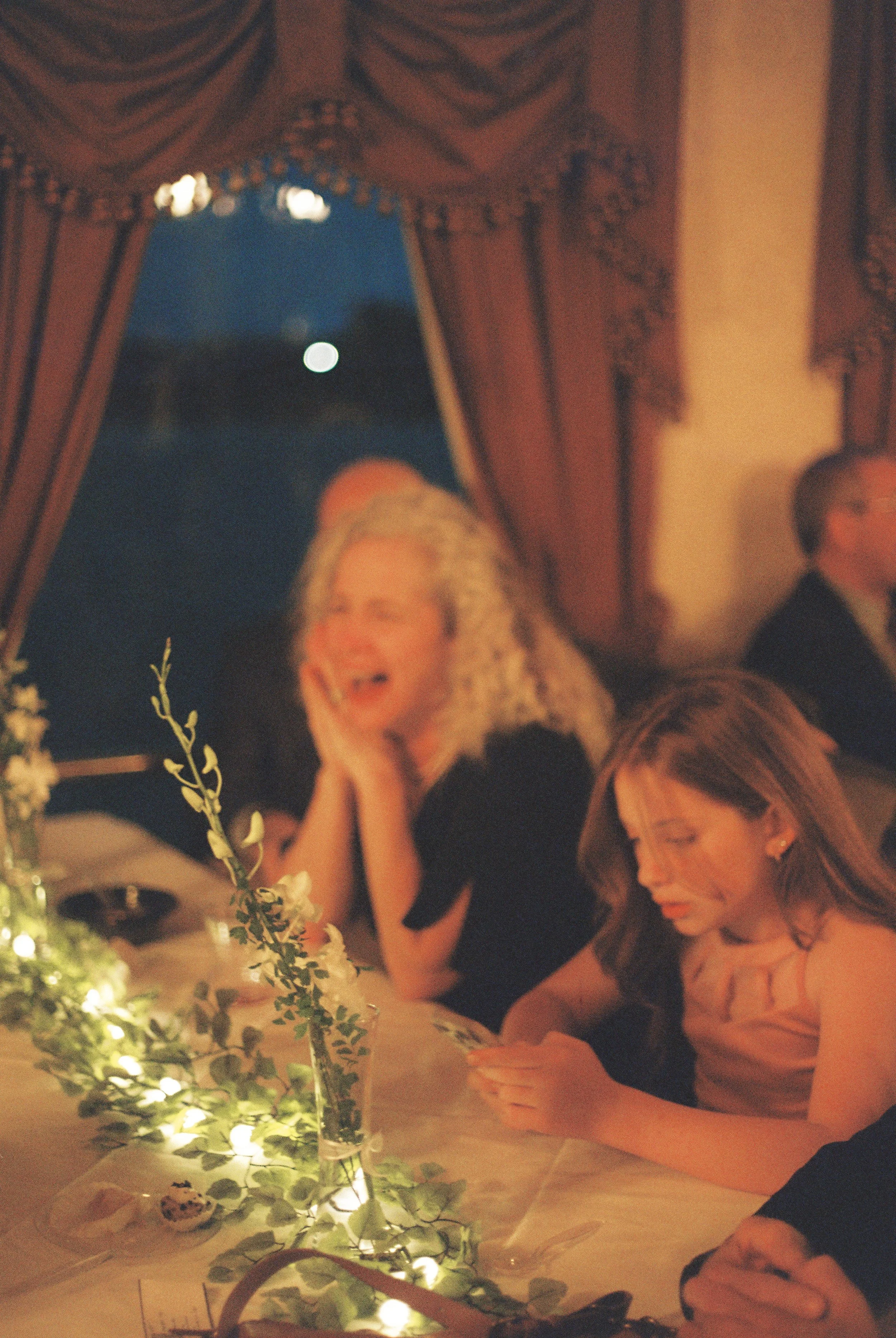 Guests seated around a candlelit dinner table decorated with greenery and soft string lights.