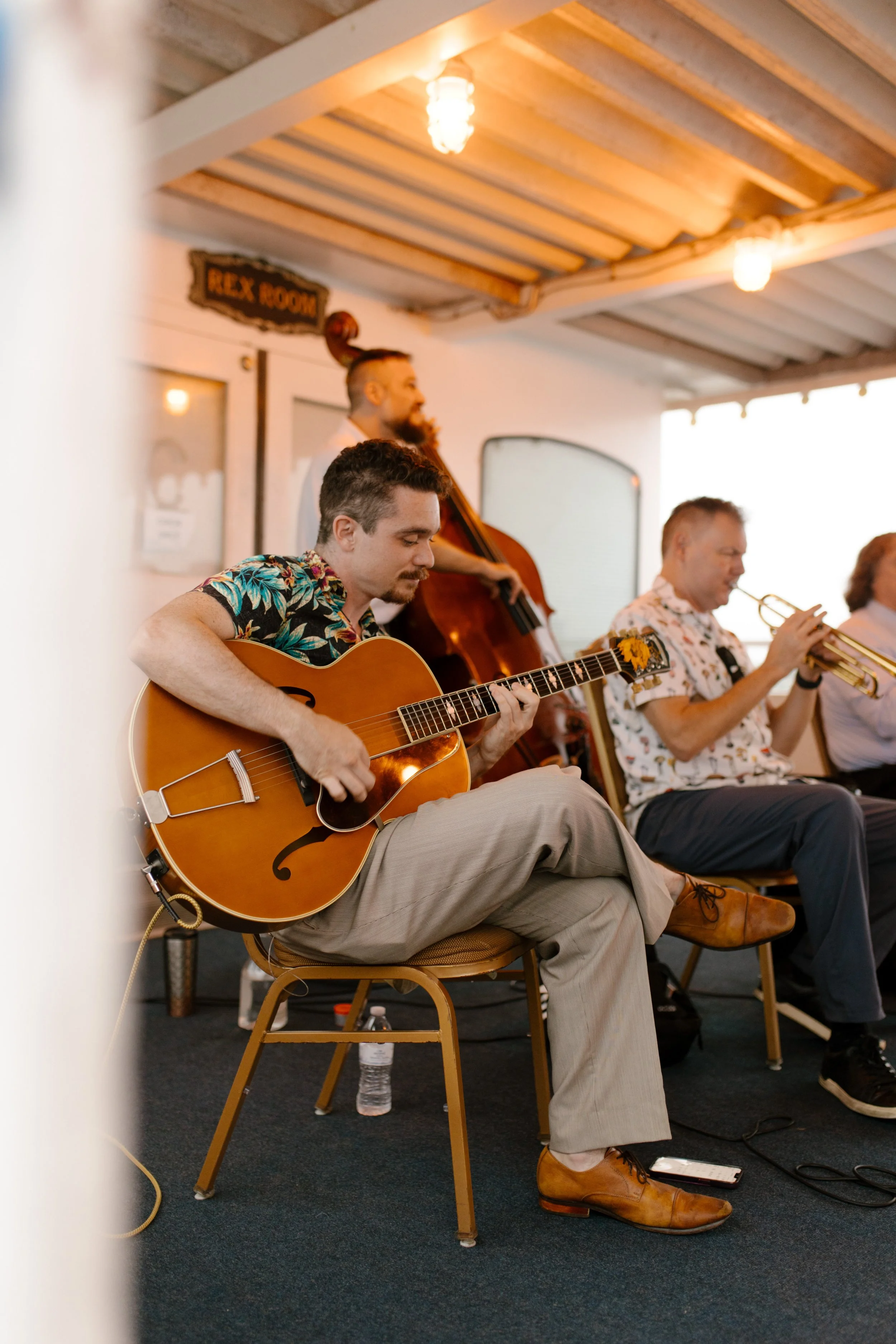 Live musicians playing guitar, upright bass, and trumpet during a wedding rehearsal dinner on a boat.