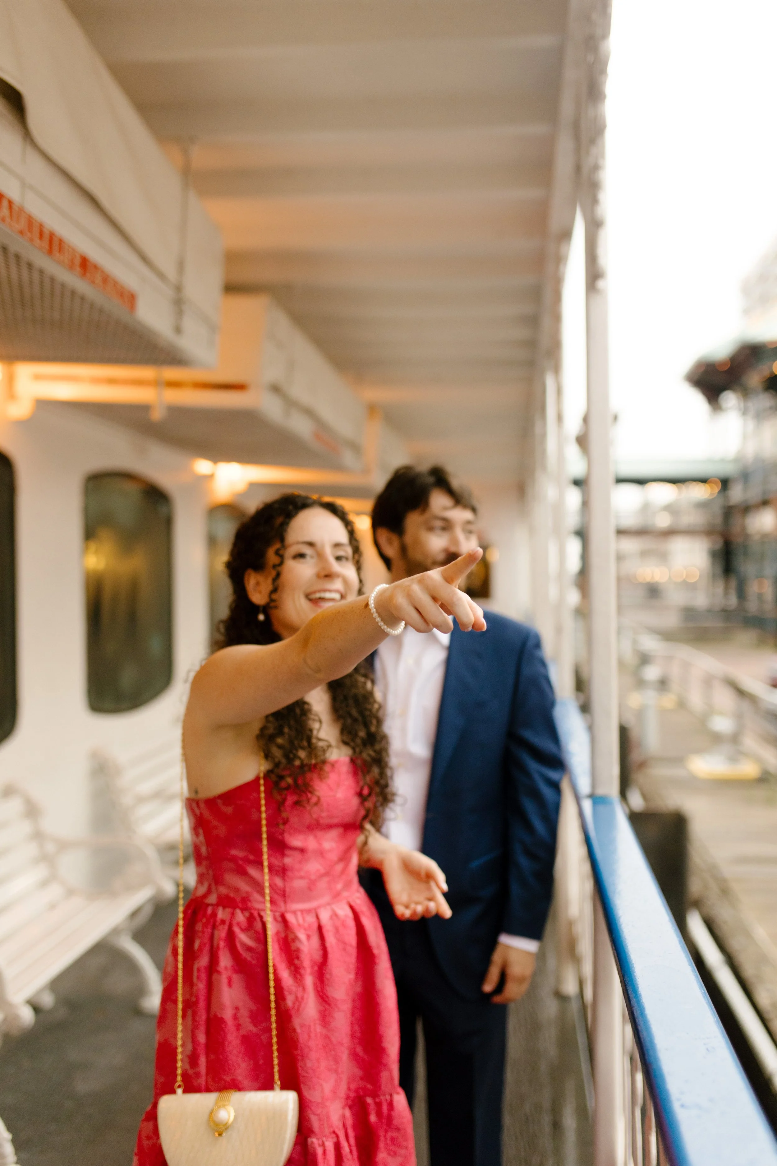 Woman in a red dress pointing toward the view while standing on the boat deck with her partner.