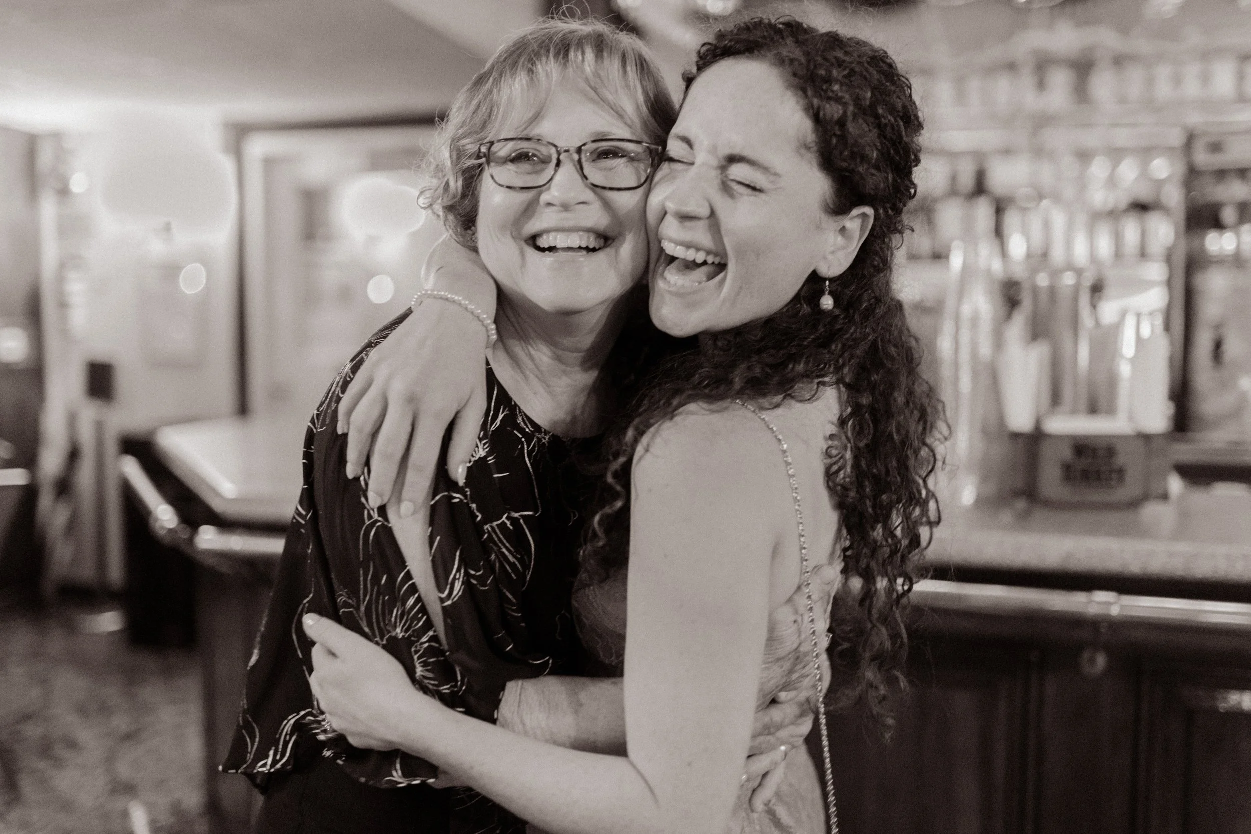 Black-and-white photo of two women hugging and laughing together during the wedding rehearsal dinner.