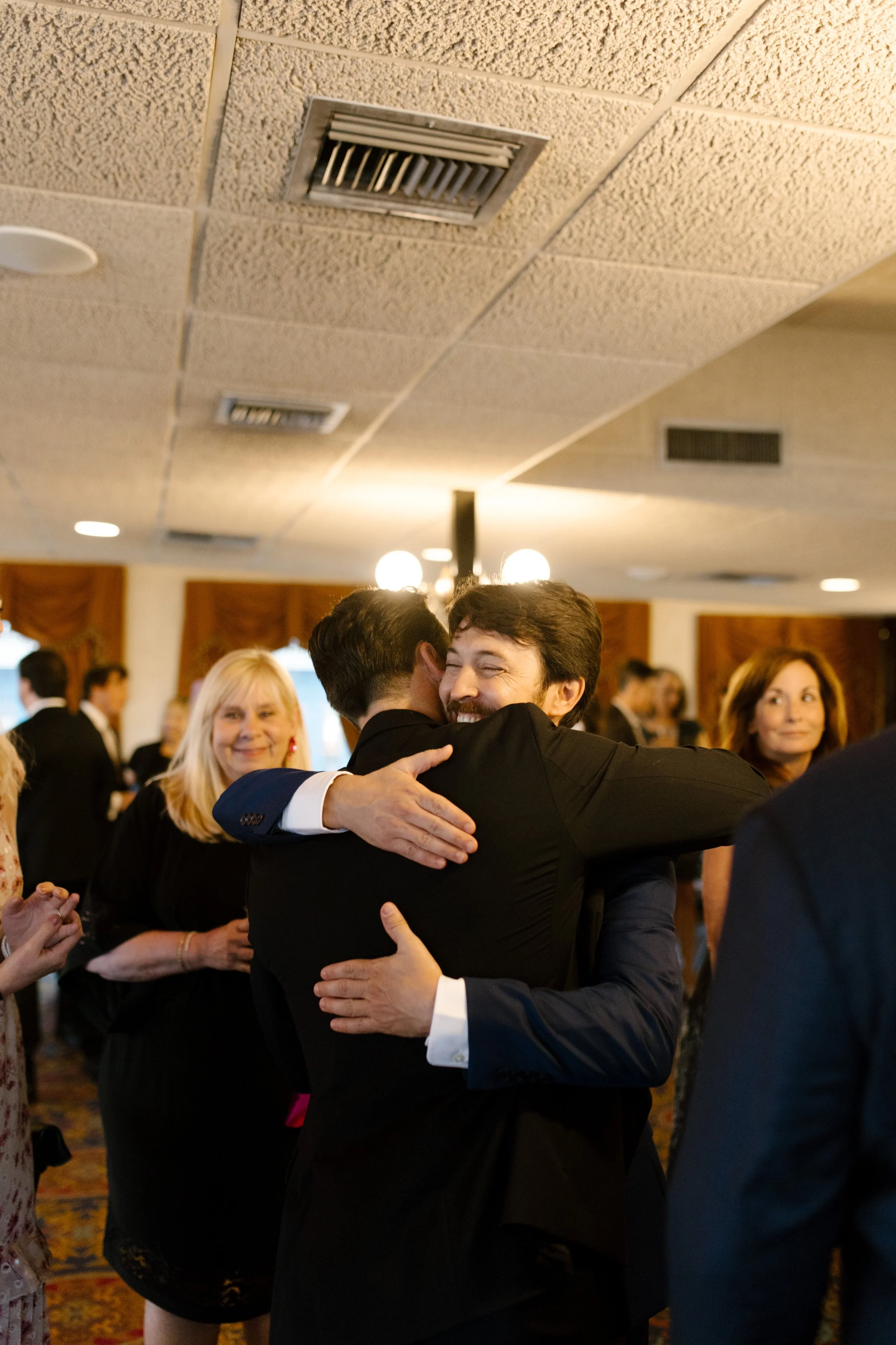 Groom-to-be warmly hugging a guest while family and friends gather around them at the celebration.
