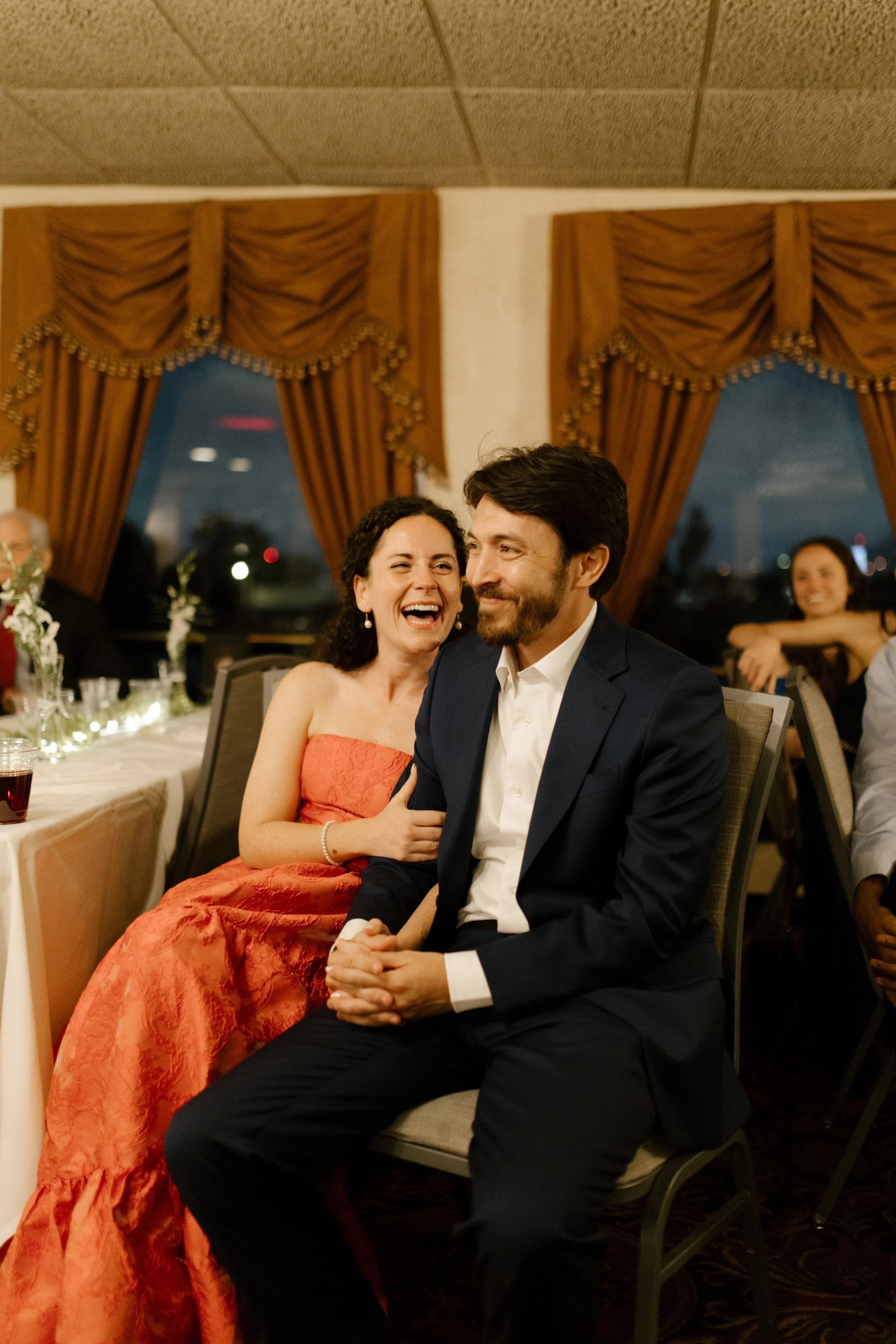 Couple laughing together while seated with their guests during the wedding rehearsal dinner.