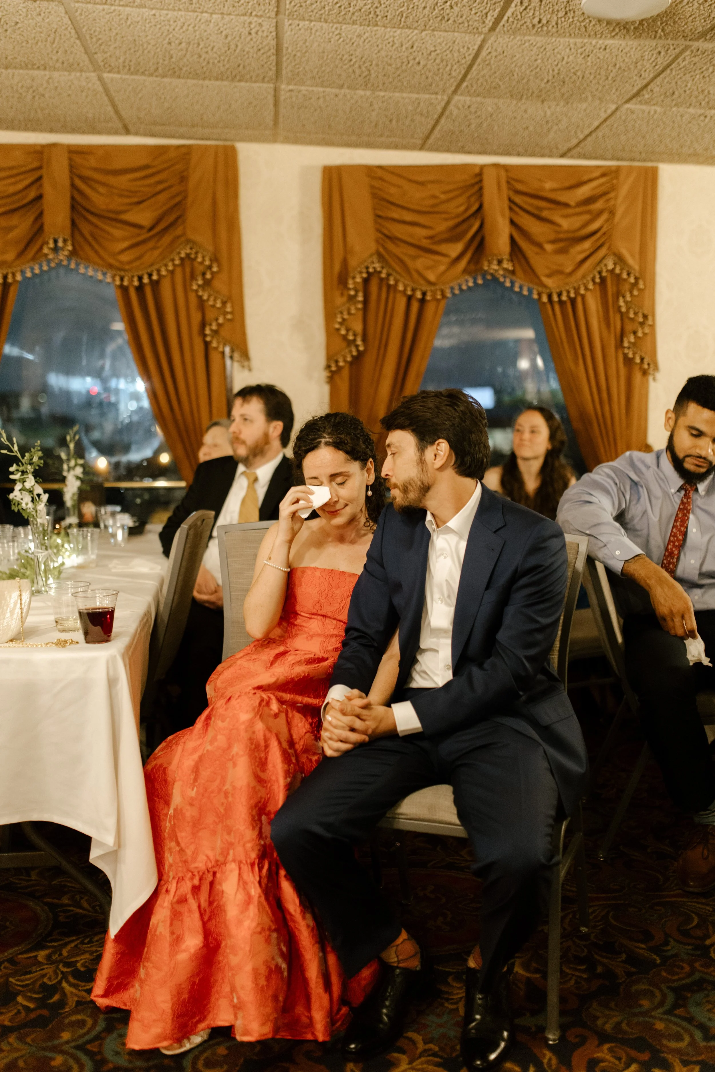 Bride-to-be wiping away tears while sitting beside her partner during their wedding rehearsal dinner.