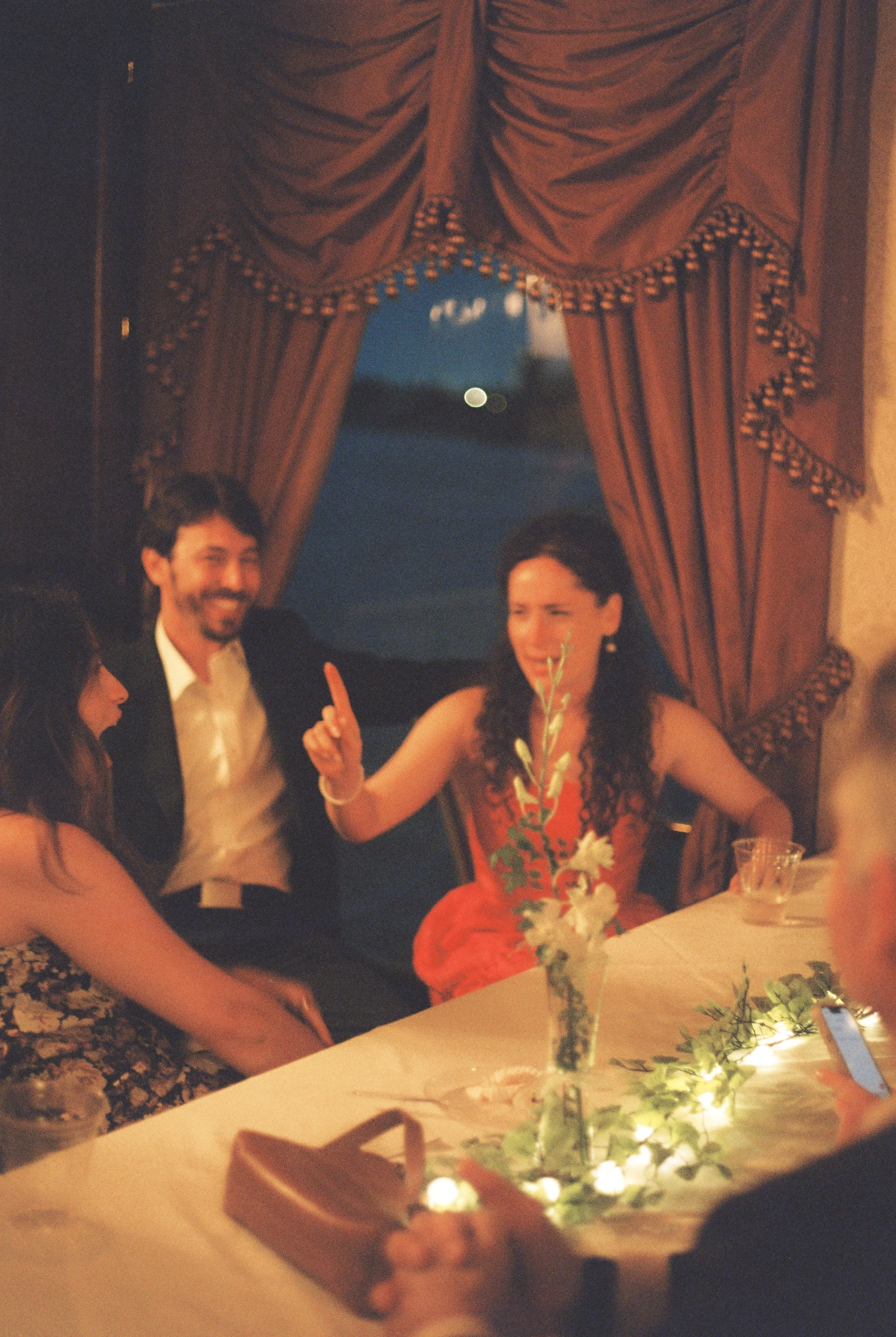 Couple sitting together at a candlelit table while chatting with guests during their rehearsal dinner celebration.