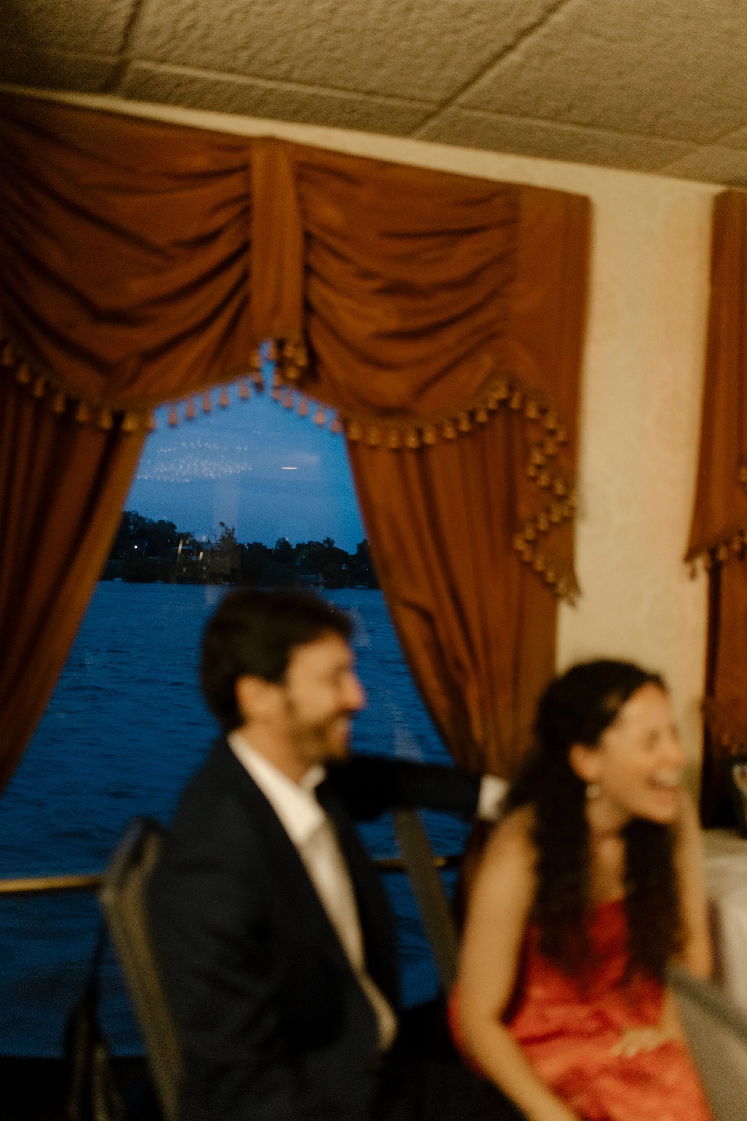 Couple laughing together beside a window overlooking the river during their wedding rehearsal dinner.