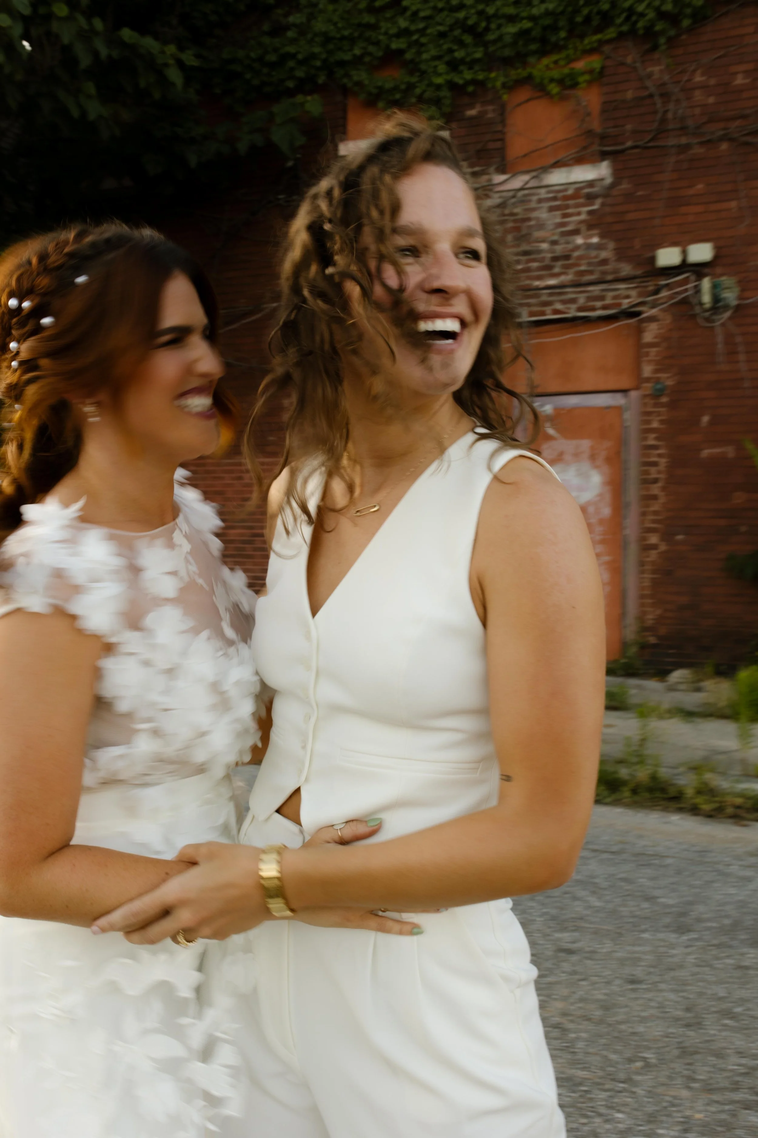 Two brides laughing together in modern white wedding outfits during their courthouse celebration