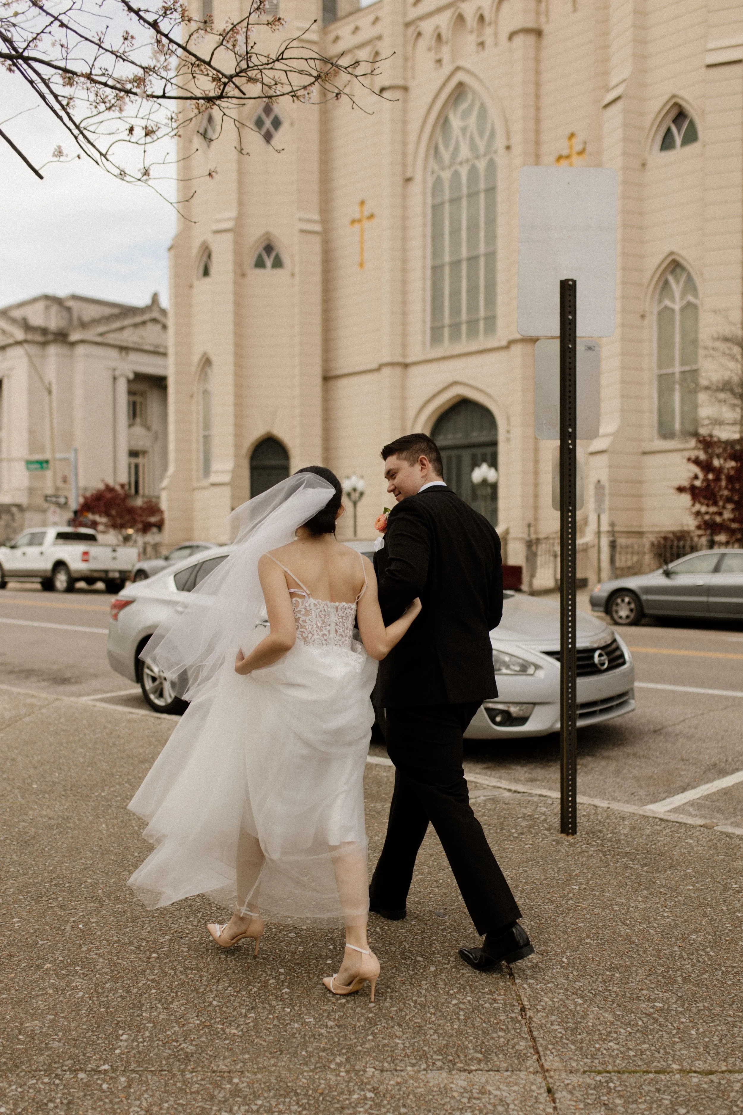 Newlyweds walking hand in hand through the city in a lace wedding dress, veil, and black tuxedo