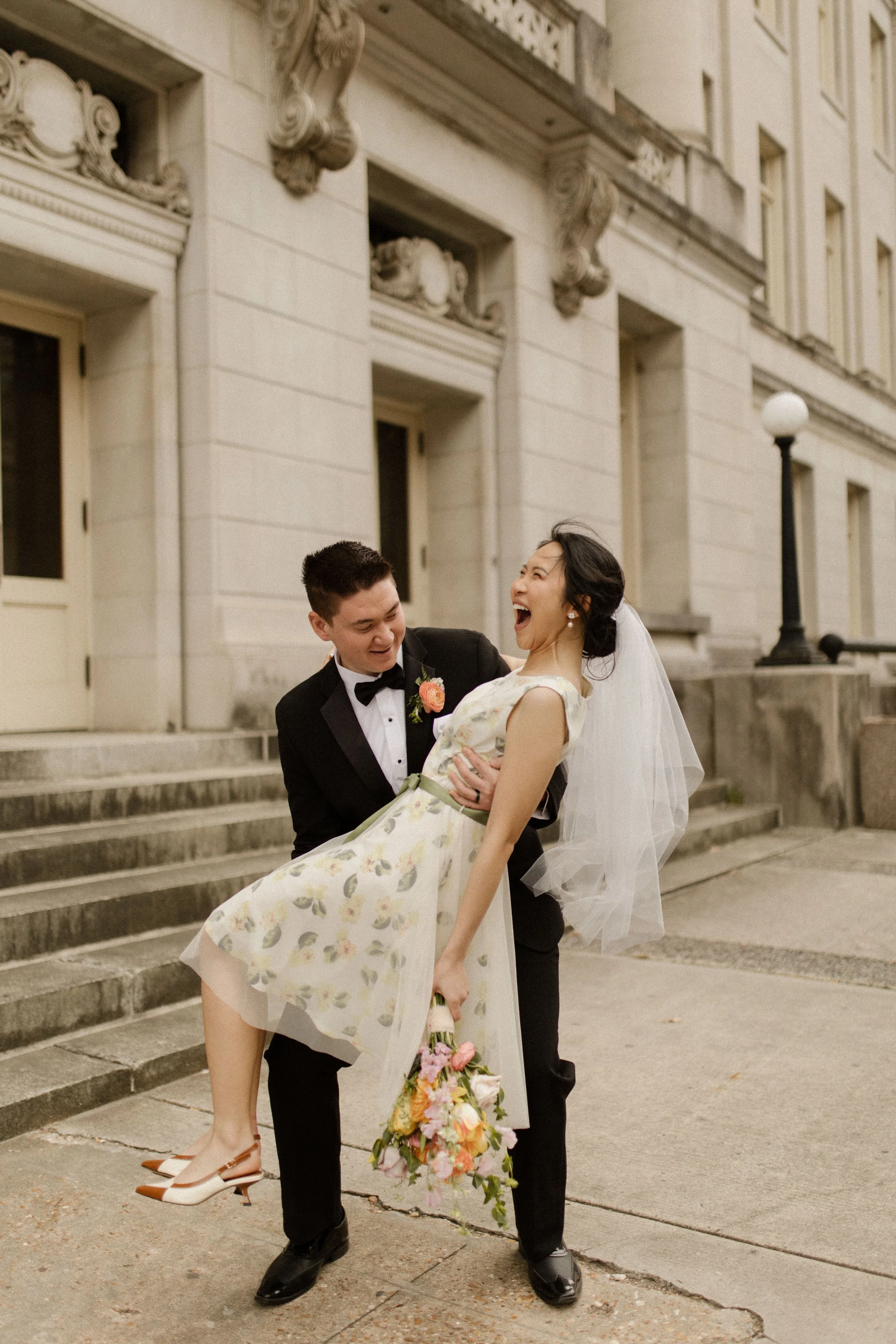 Newlyweds laughing outside the courthouse with the bride wearing a floral wedding dress, veil, and colorful bouquet