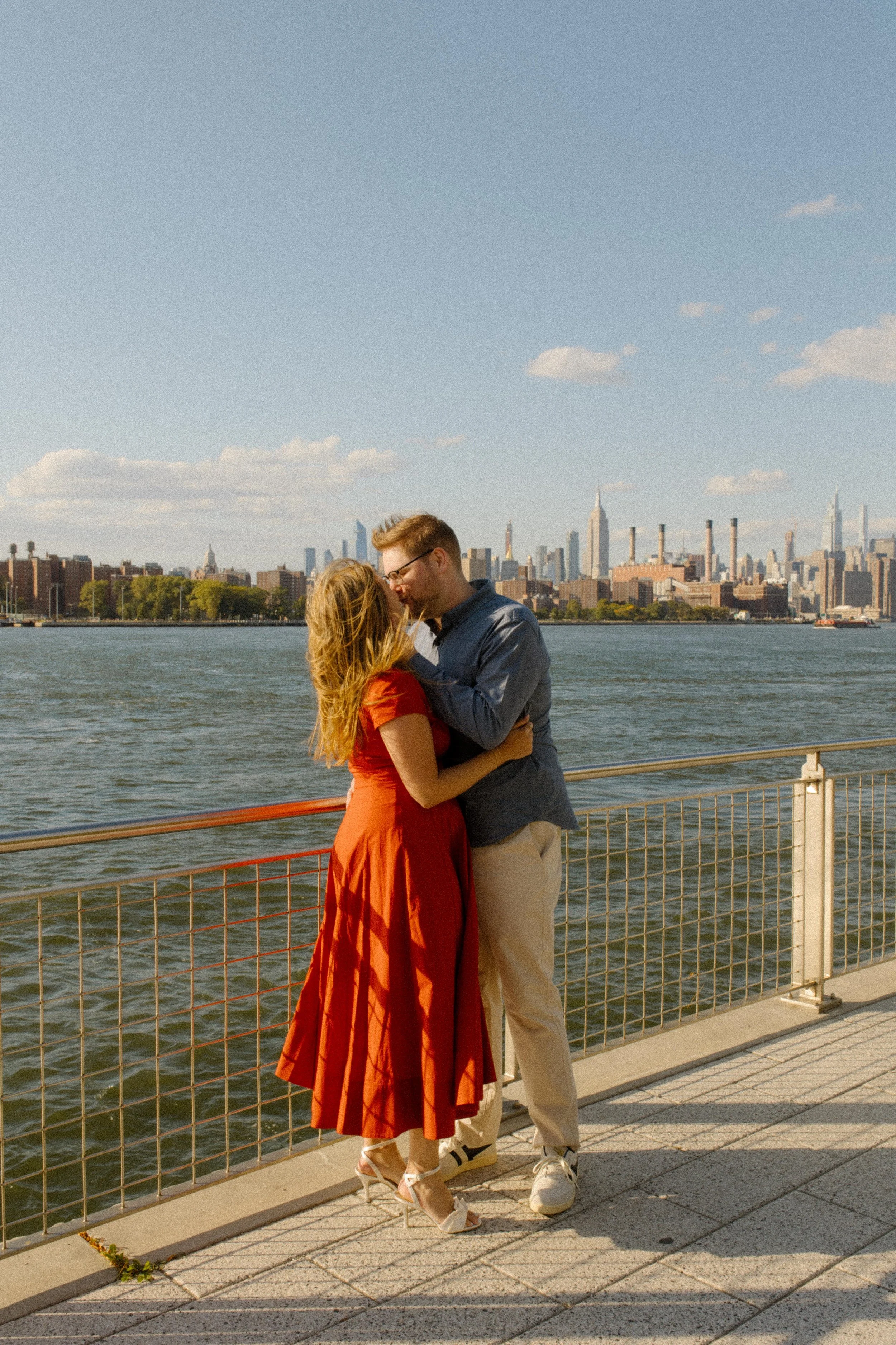Couple embracing along the East River waterfront with the Manhattan skyline glowing behind them at sunset.