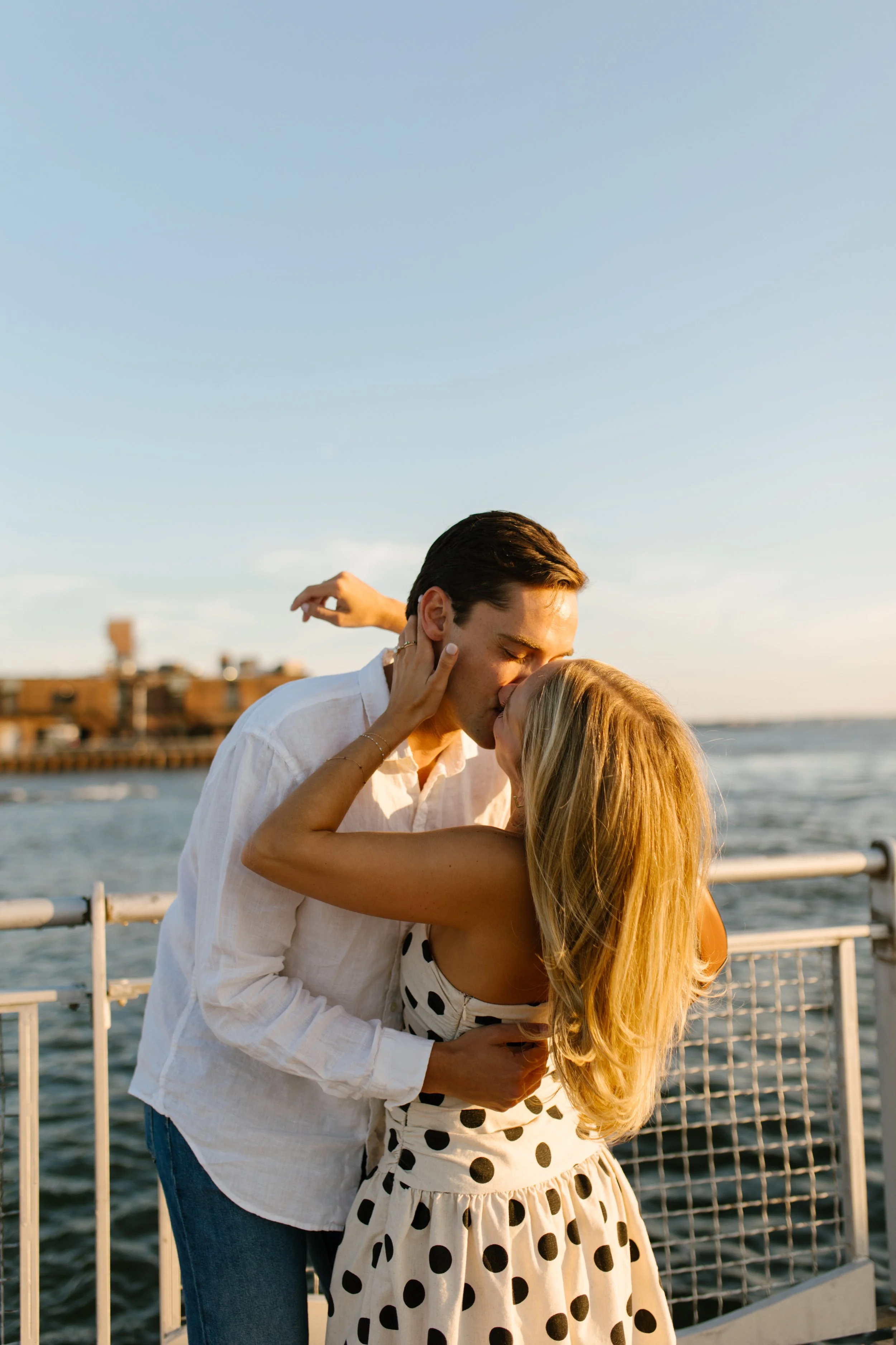 Couple kiss beside the waterfront in warm golden-hour light during their New York engagement session.