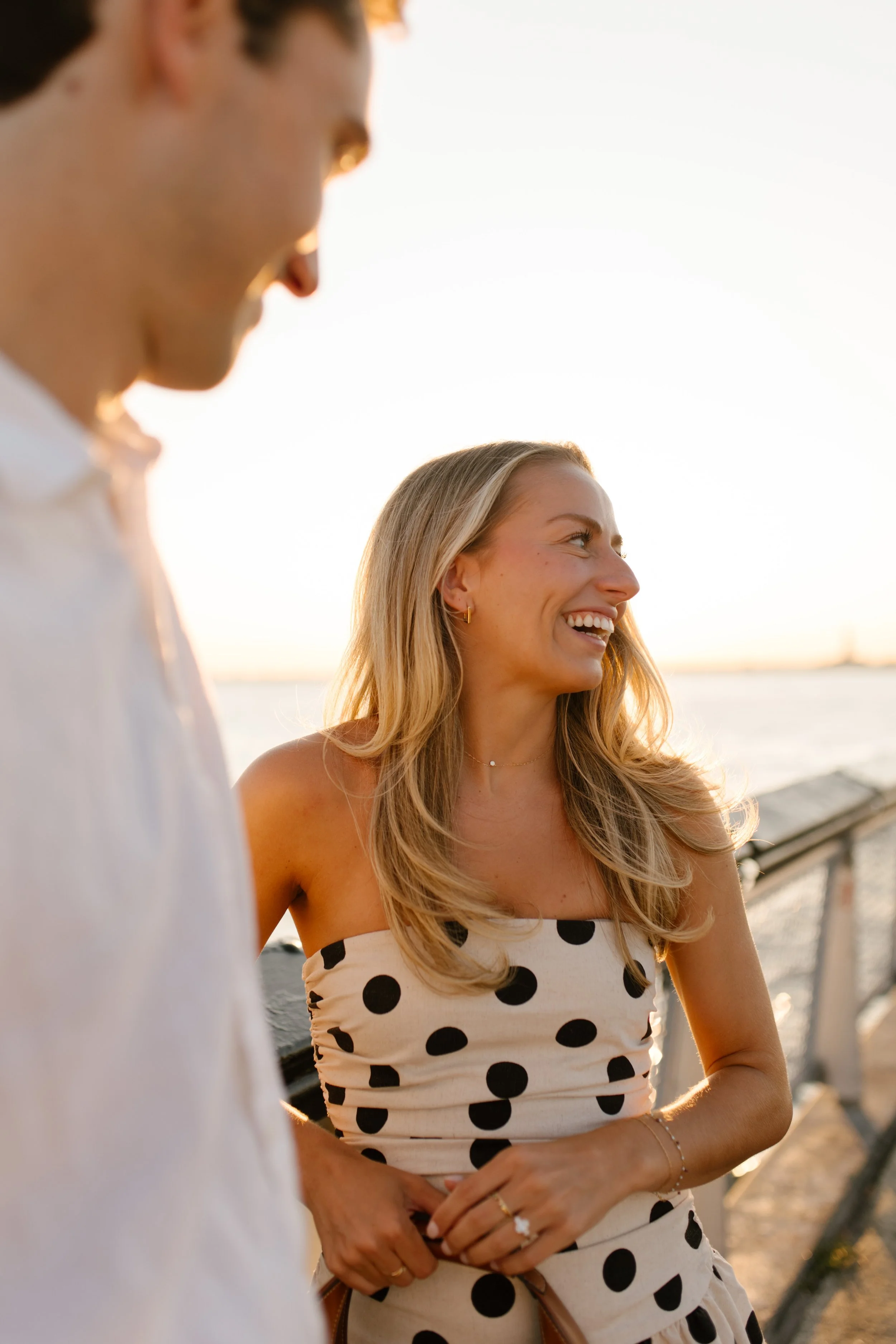 Woman in a polka-dot dress smiles at her partner during a golden-hour waterfront engagement session.