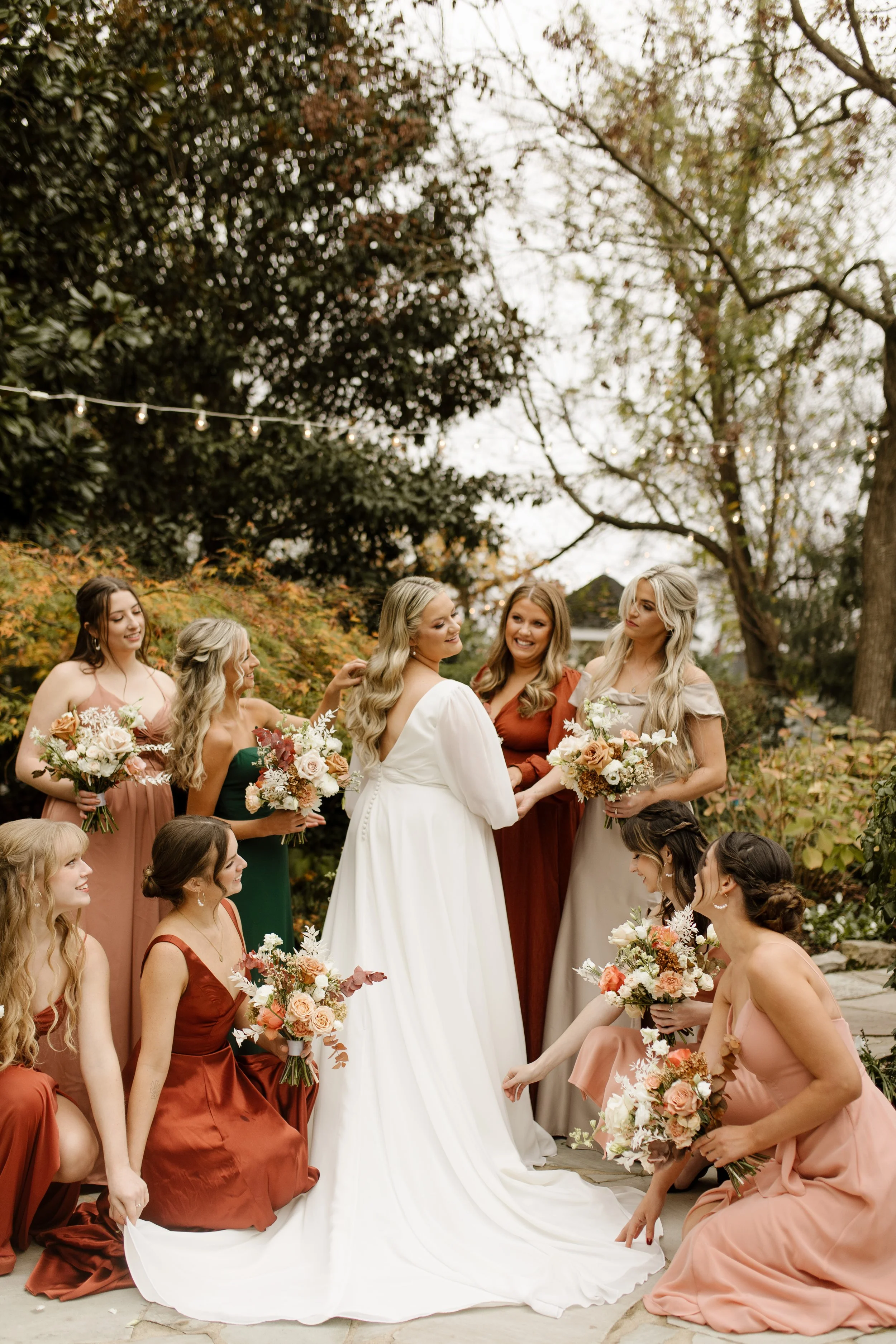 Bride surrounded by bridesmaids in rust, green, and blush dresses holding autumn bouquets at an outdoor fall wedding.