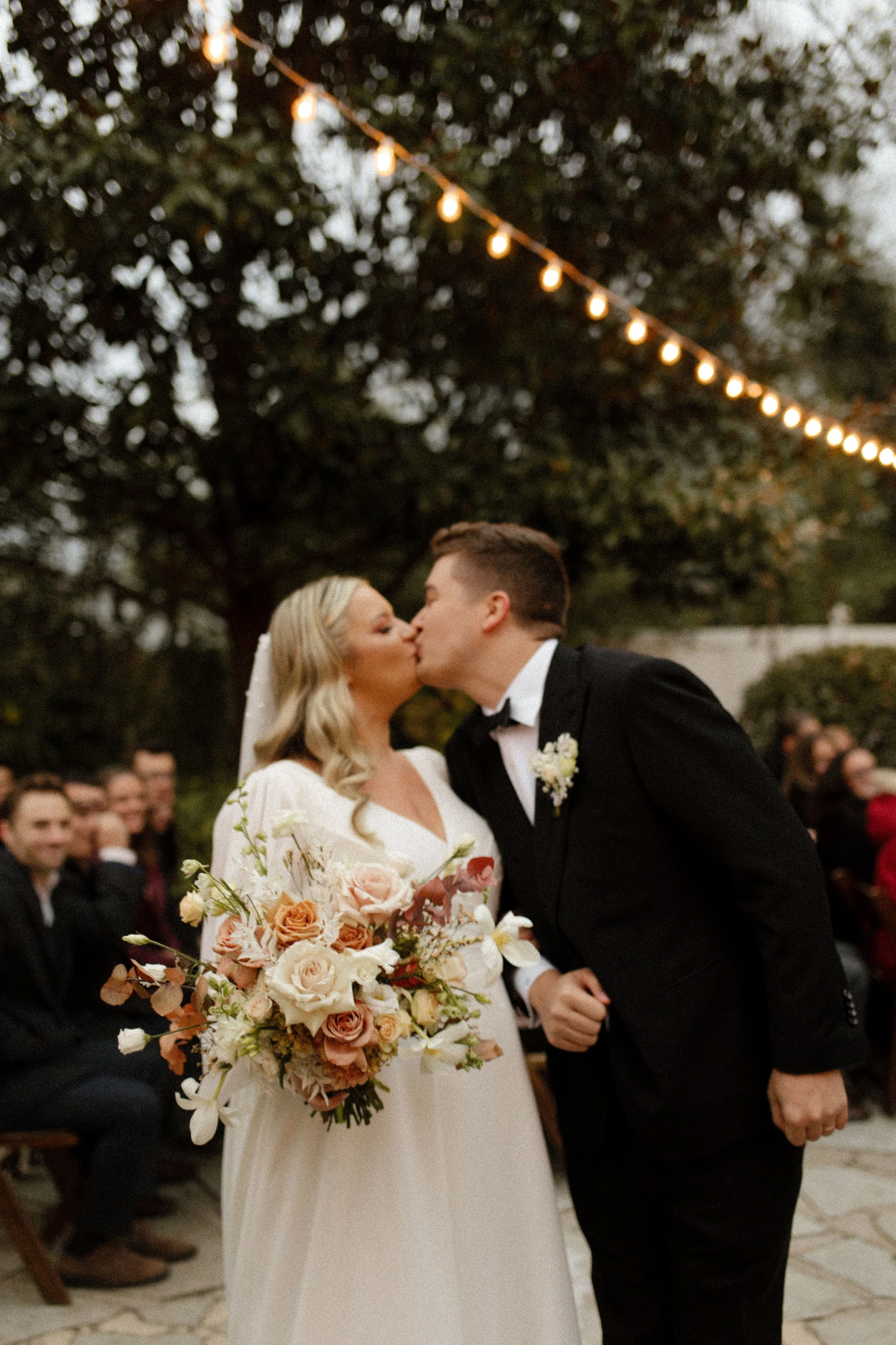 Newlyweds sharing a kiss after their outdoor ceremony beneath glowing string lights and surrounded by wedding guests.
