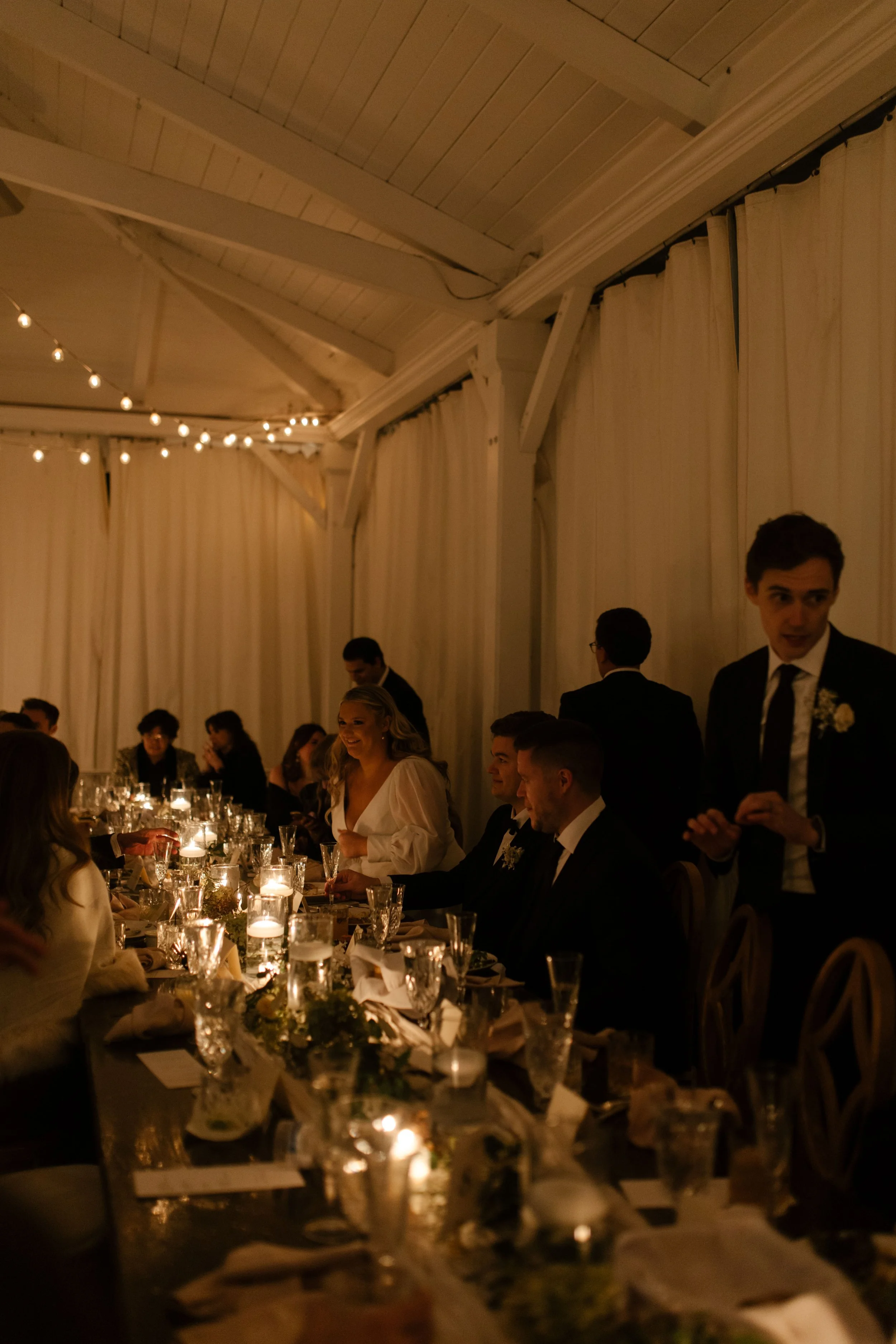 Candlelit fall wedding reception with guests gathered around long dinner tables beneath warm string lights.