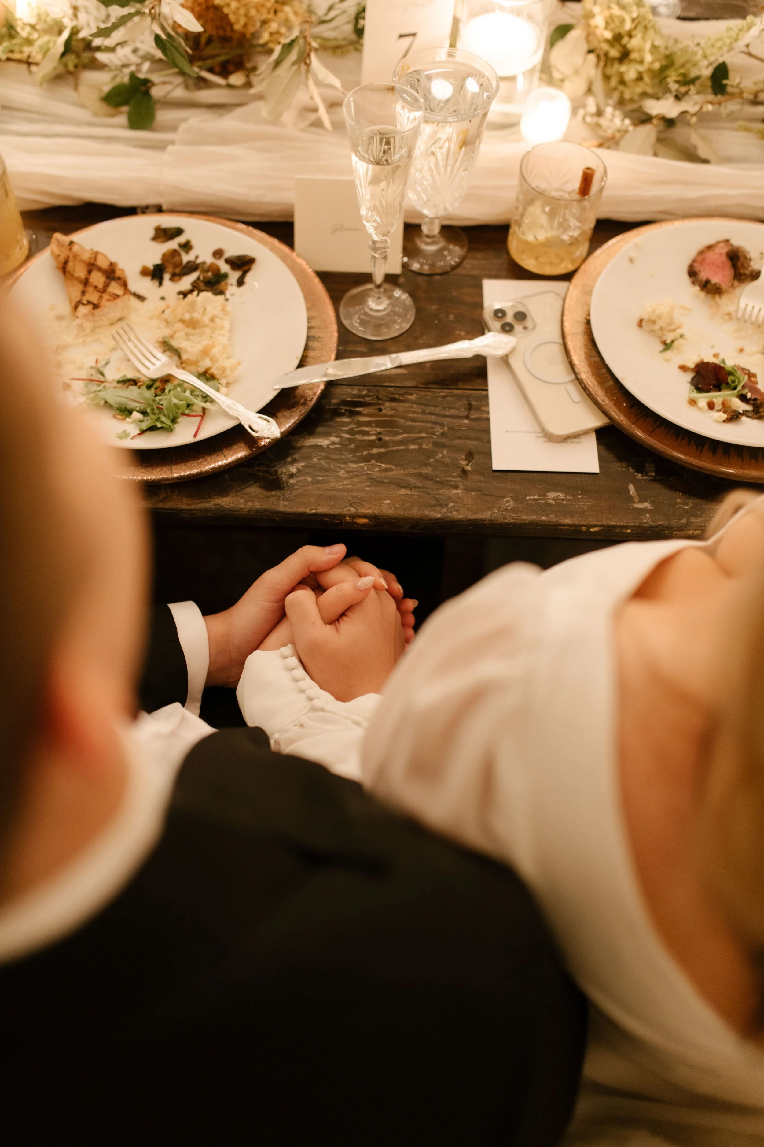 Candid overhead photo of the newlyweds holding hands together beneath the dinner table during their wedding reception.