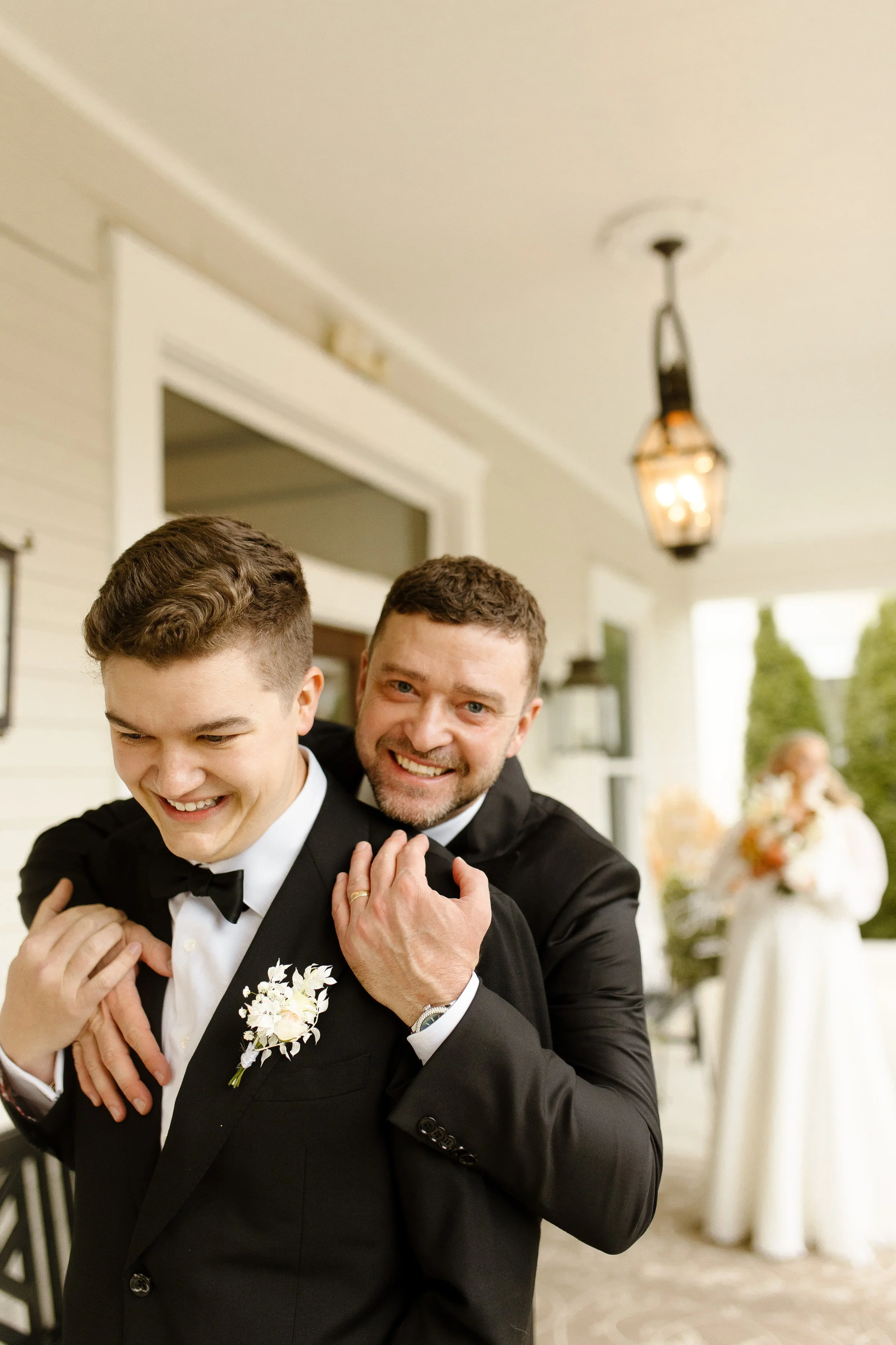 Groom laughing with a groomsman on the porch while the bride stands in the background holding her bouquet.