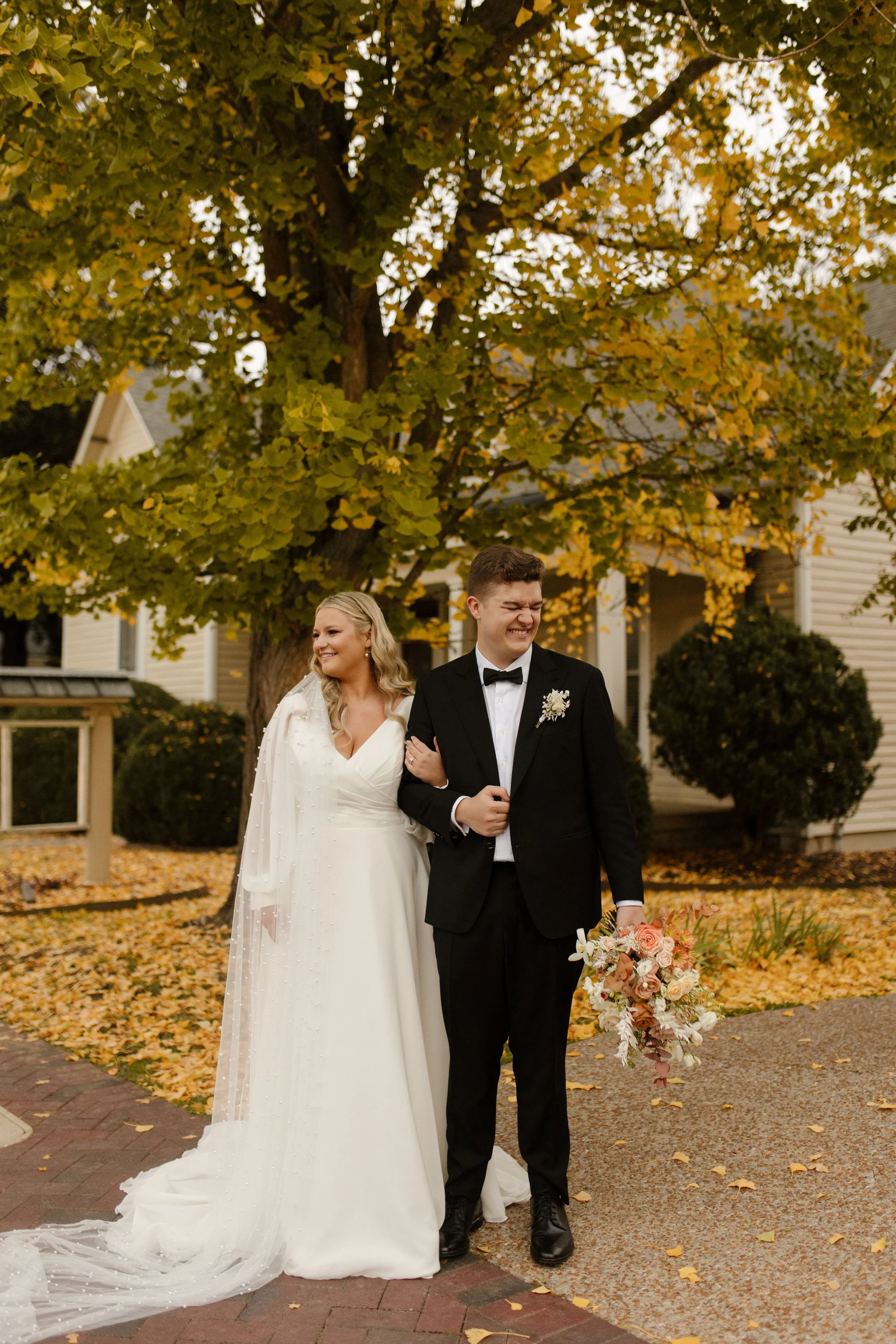 Bride and groom posing beneath golden autumn trees with a peach and cream bouquet at their fall wedding.