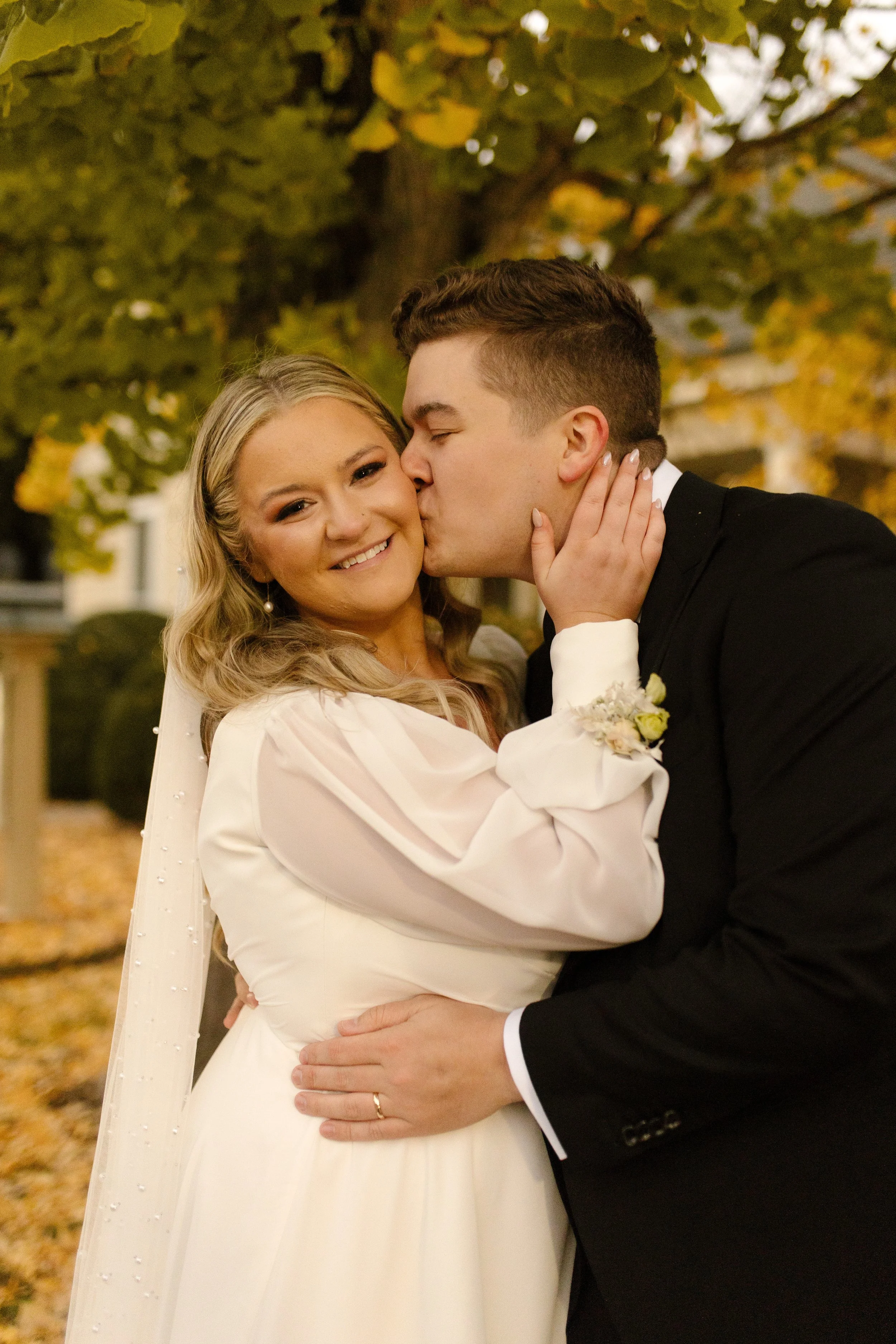 Groom kissing the bride on the cheek beneath golden autumn leaves during their outdoor wedding portraits.