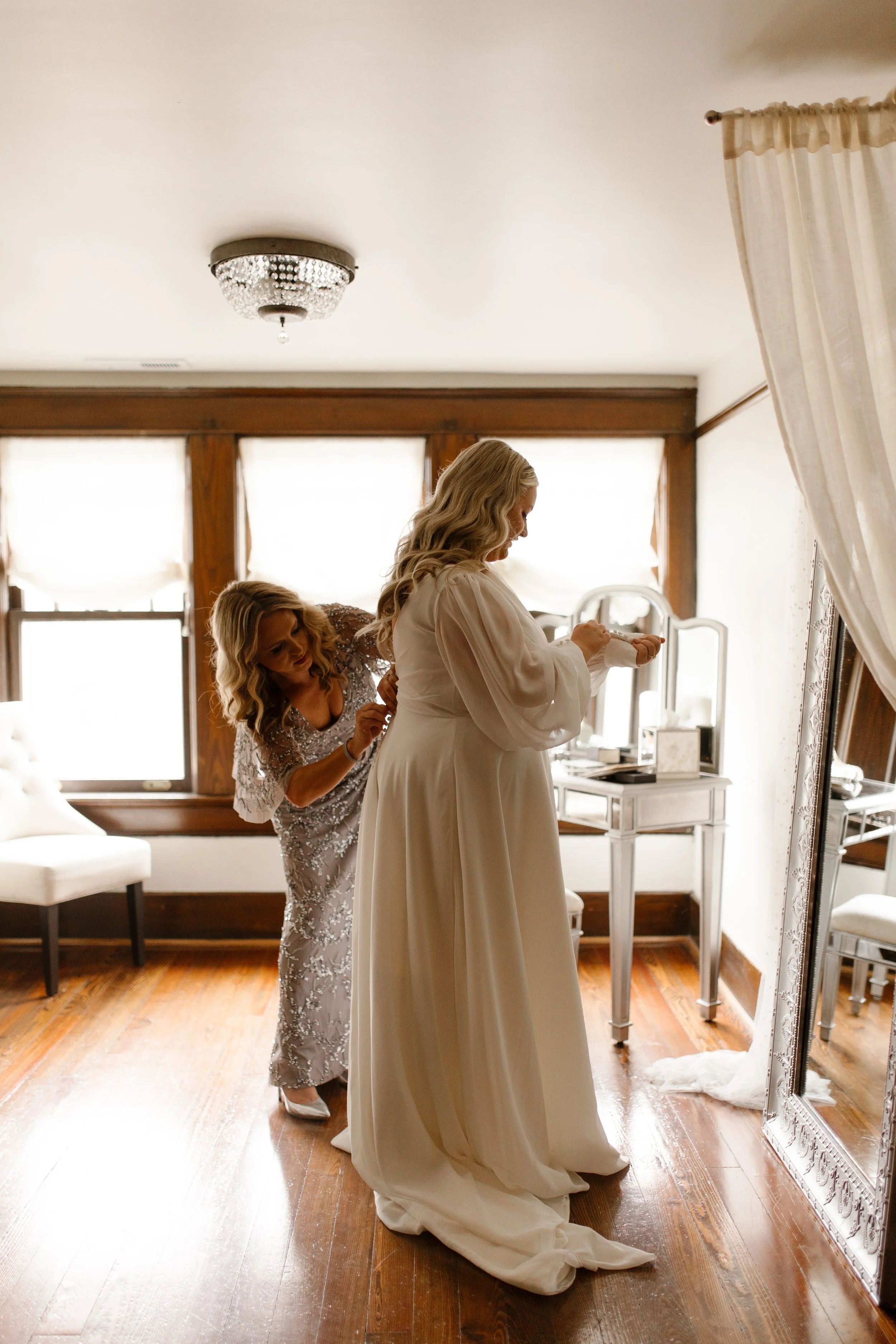 Bride getting buttoned into her long-sleeved wedding dress beside a full-length mirror before the ceremony.