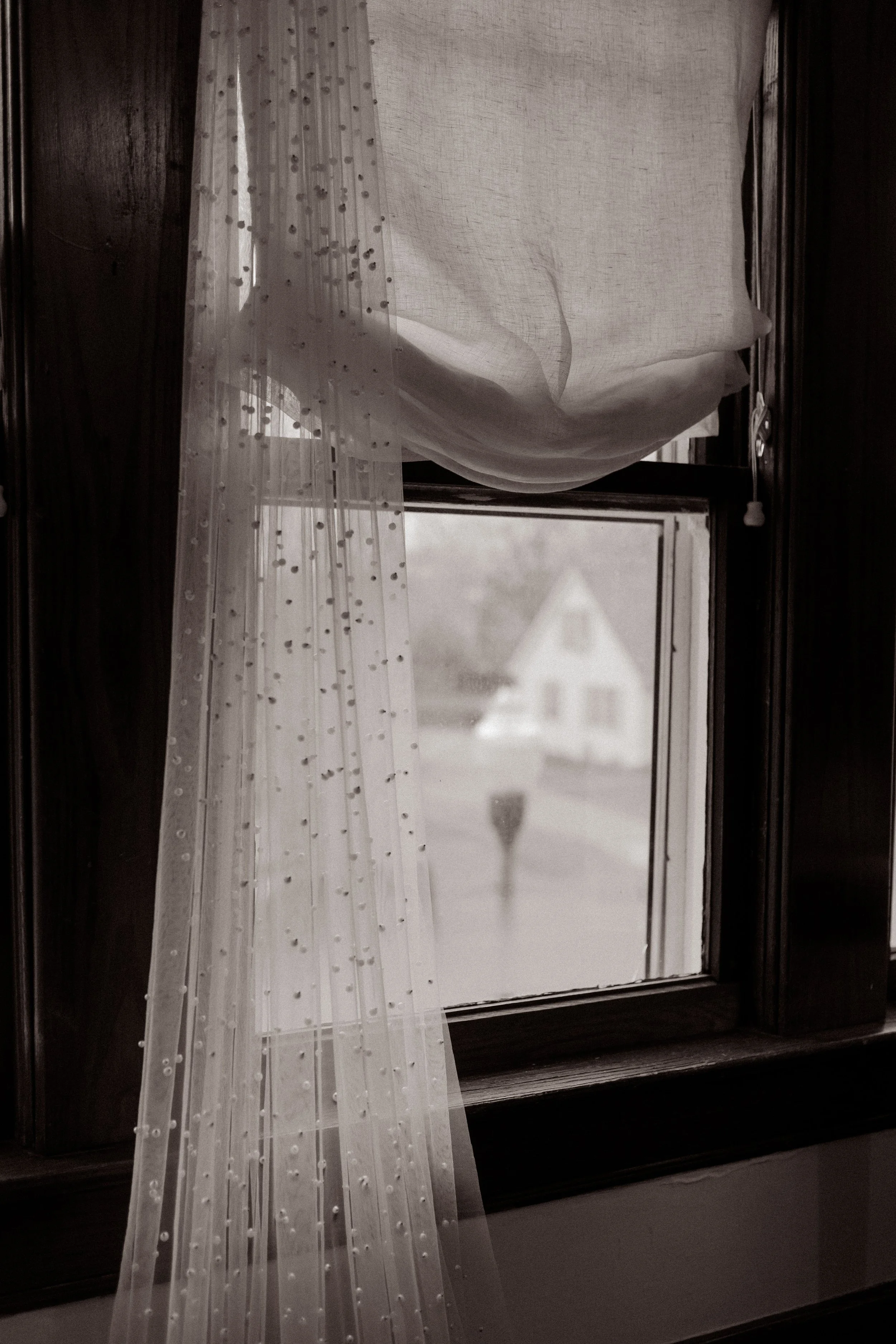 Black-and-white detail photo of the bride’s pearl-dotted wedding veil hanging beside a window.