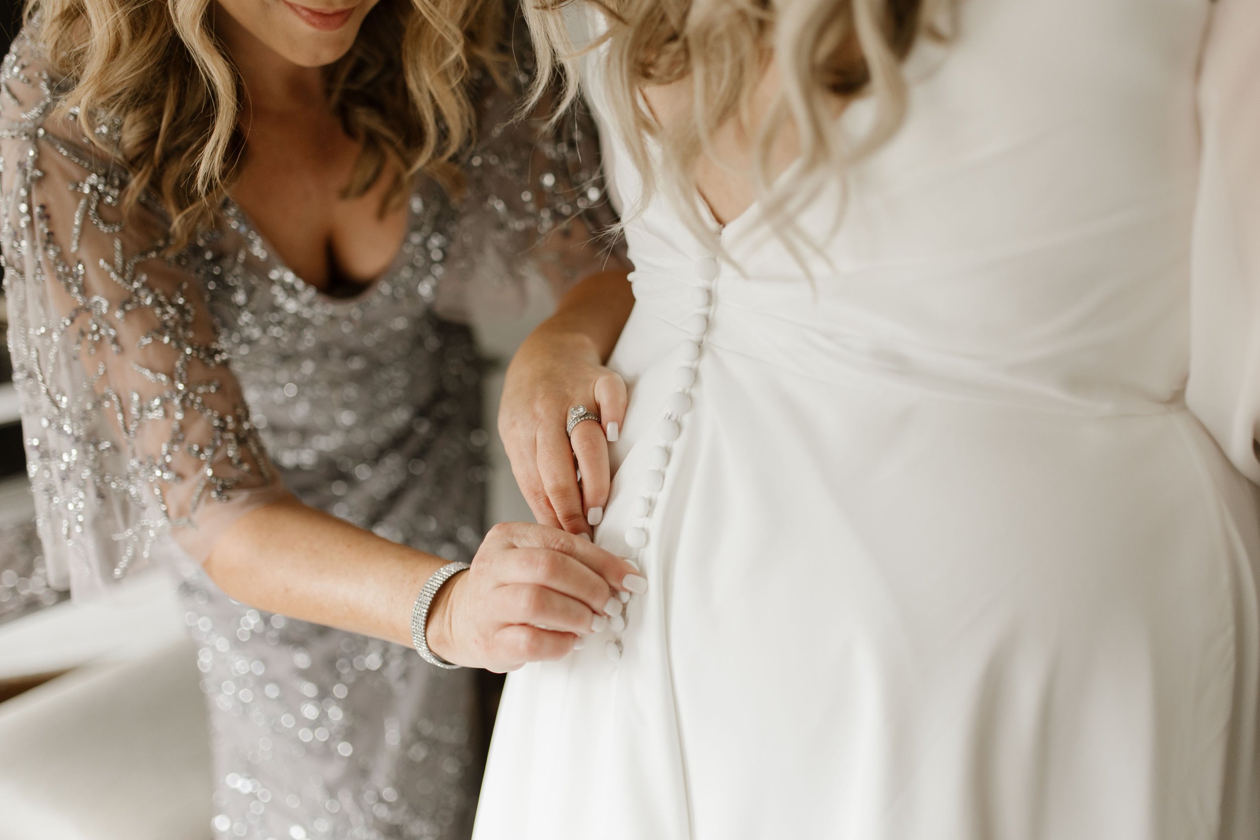 Close-up of the bride’s long-sleeved wedding dress being buttoned with her engagement ring visible.
