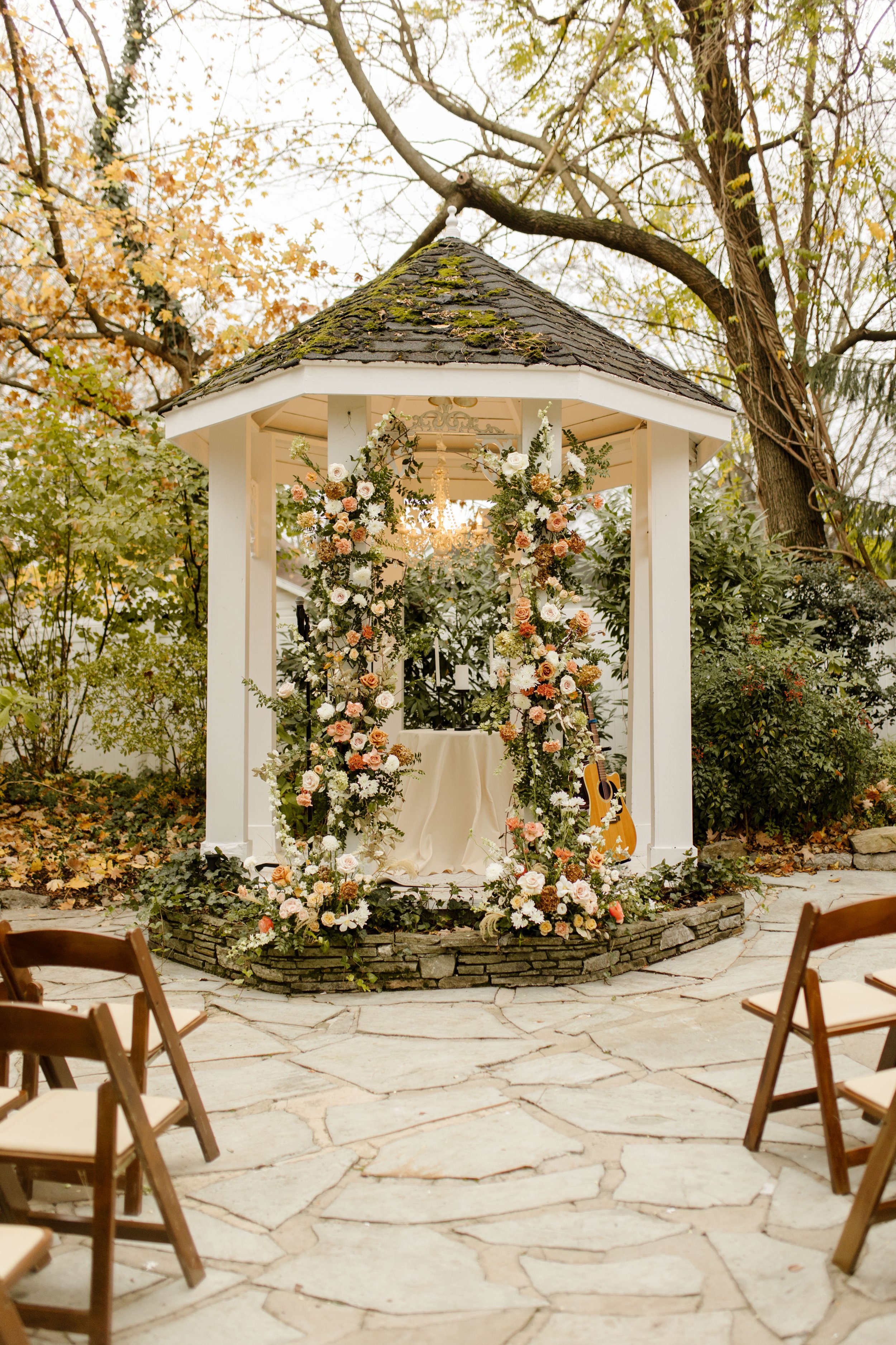 Outdoor fall wedding ceremony gazebo covered in peach, cream, and white flowers surrounded by autumn trees.