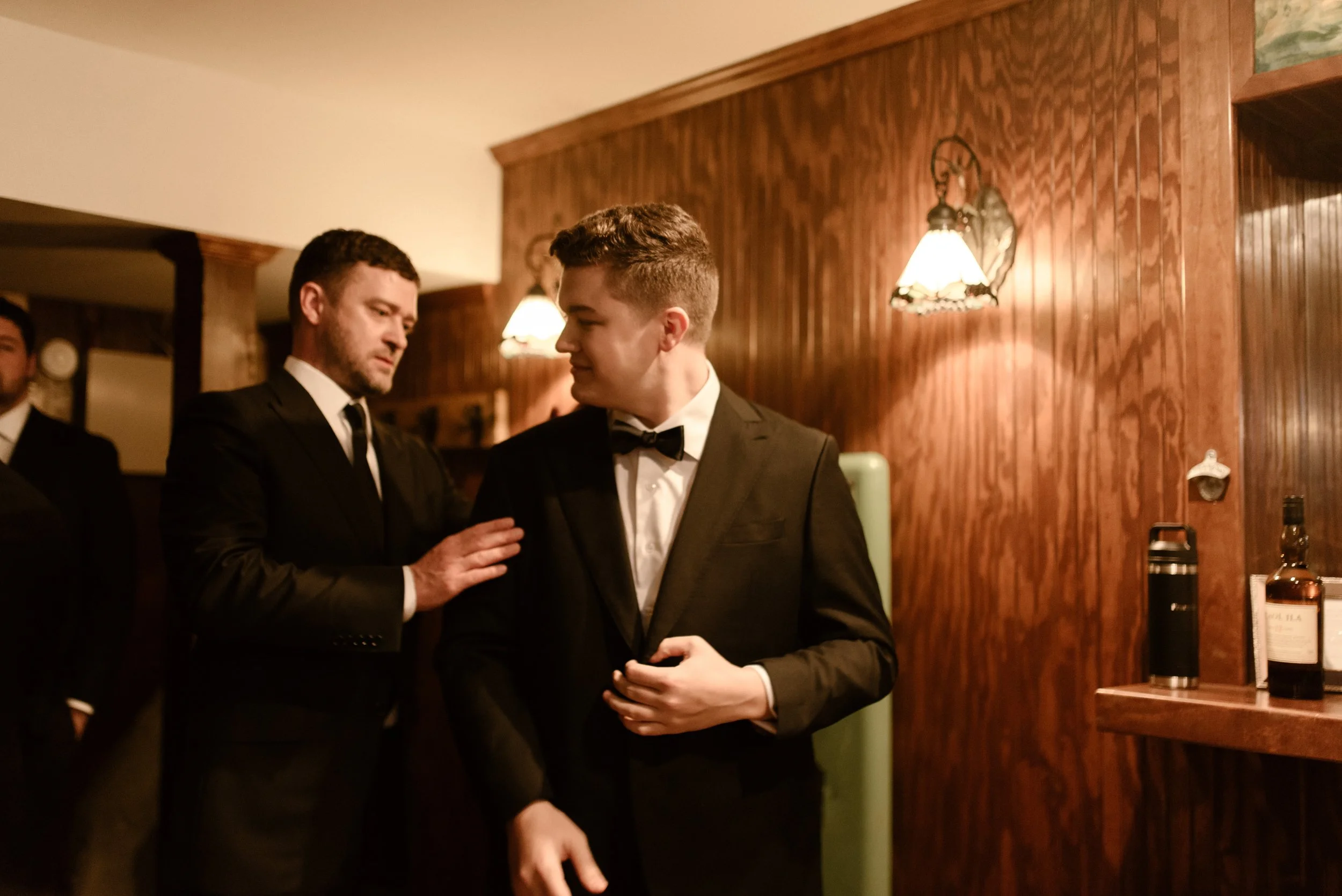 Groom in a classic black tuxedo sharing a candid moment with a groomsman while getting ready for his fall wedding.
