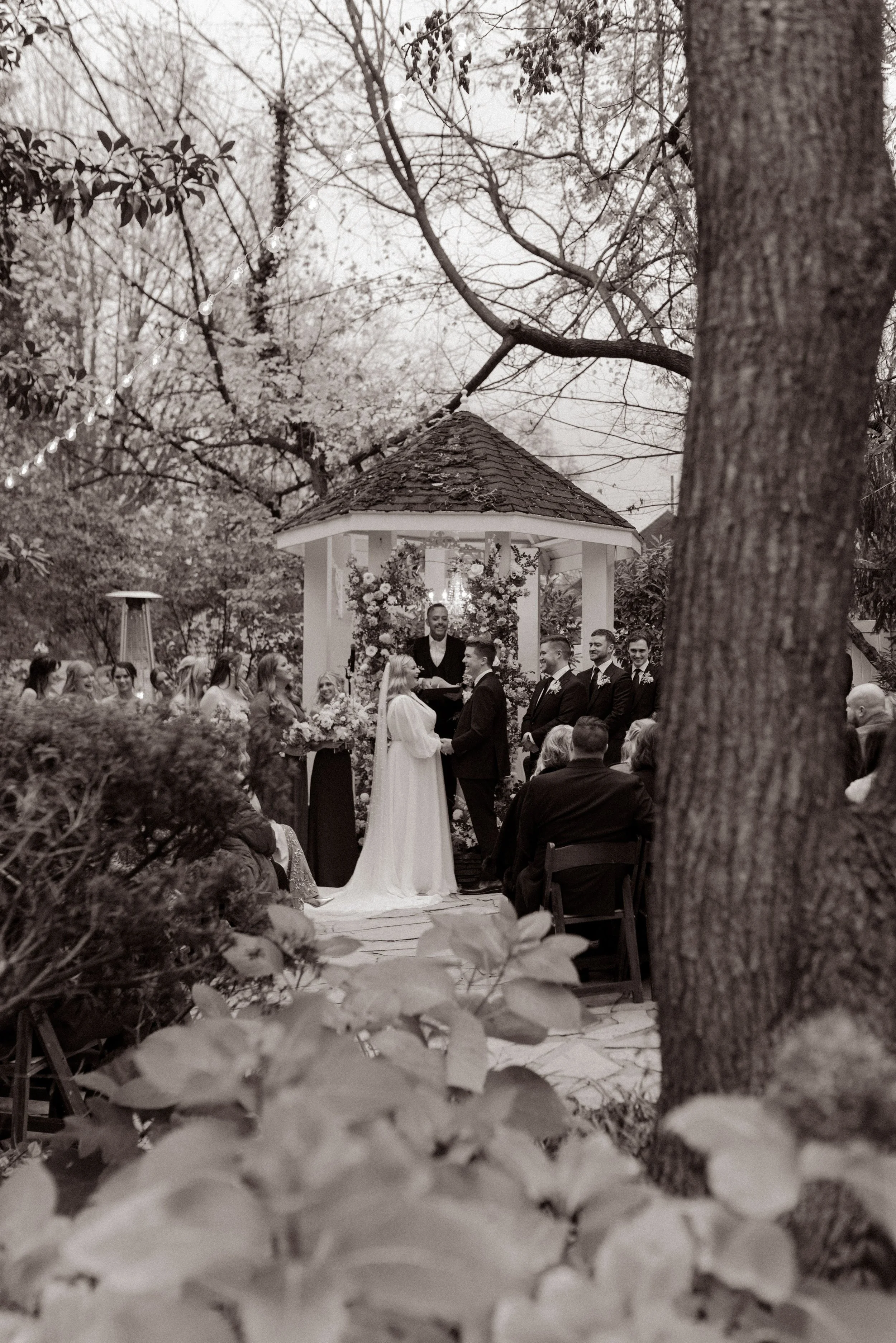 Black-and-white photo of the bride and groom exchanging vows at a flower-covered outdoor gazebo.