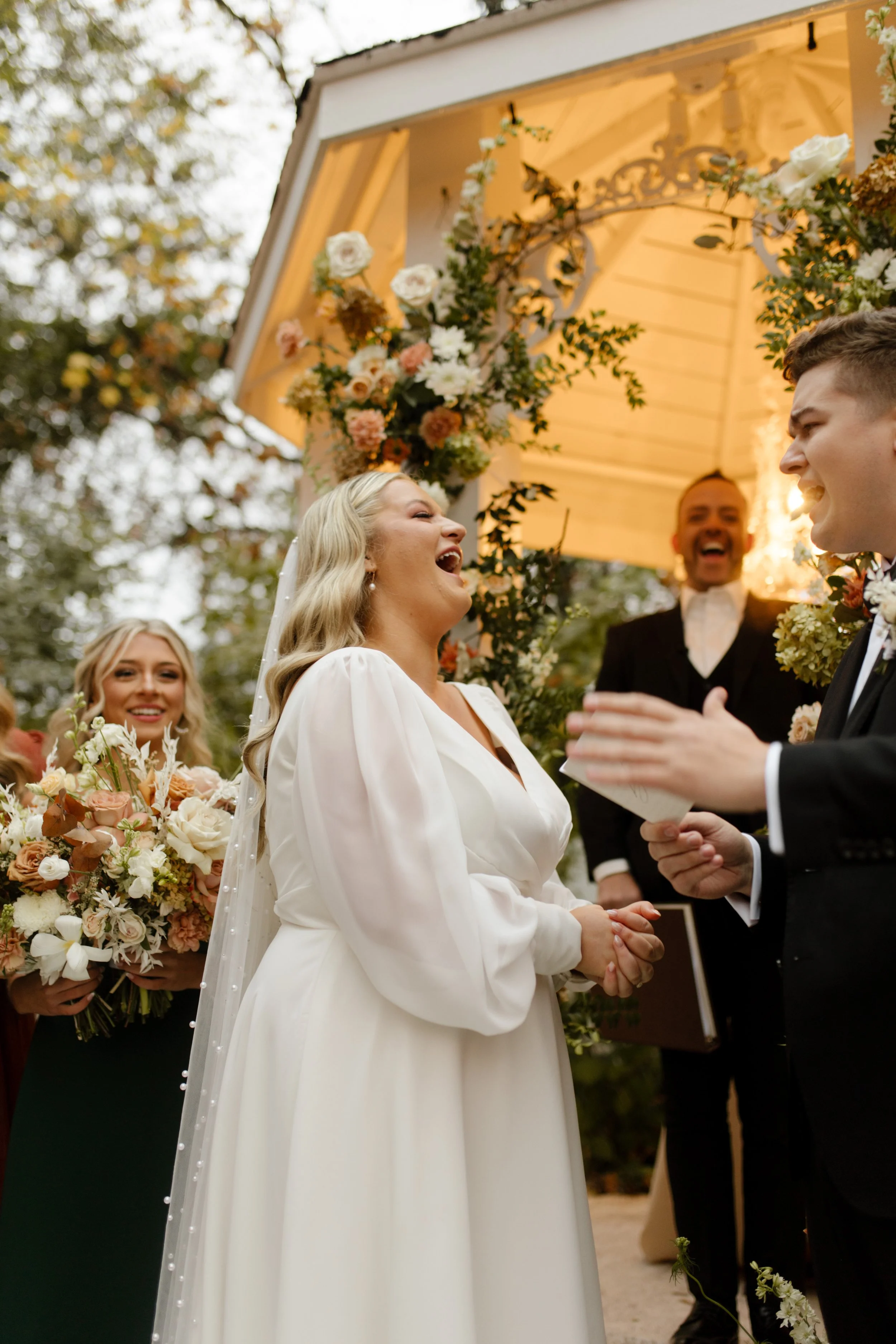 Bride laughing with the groom during their outdoor ceremony with warm autumn florals behind them.
