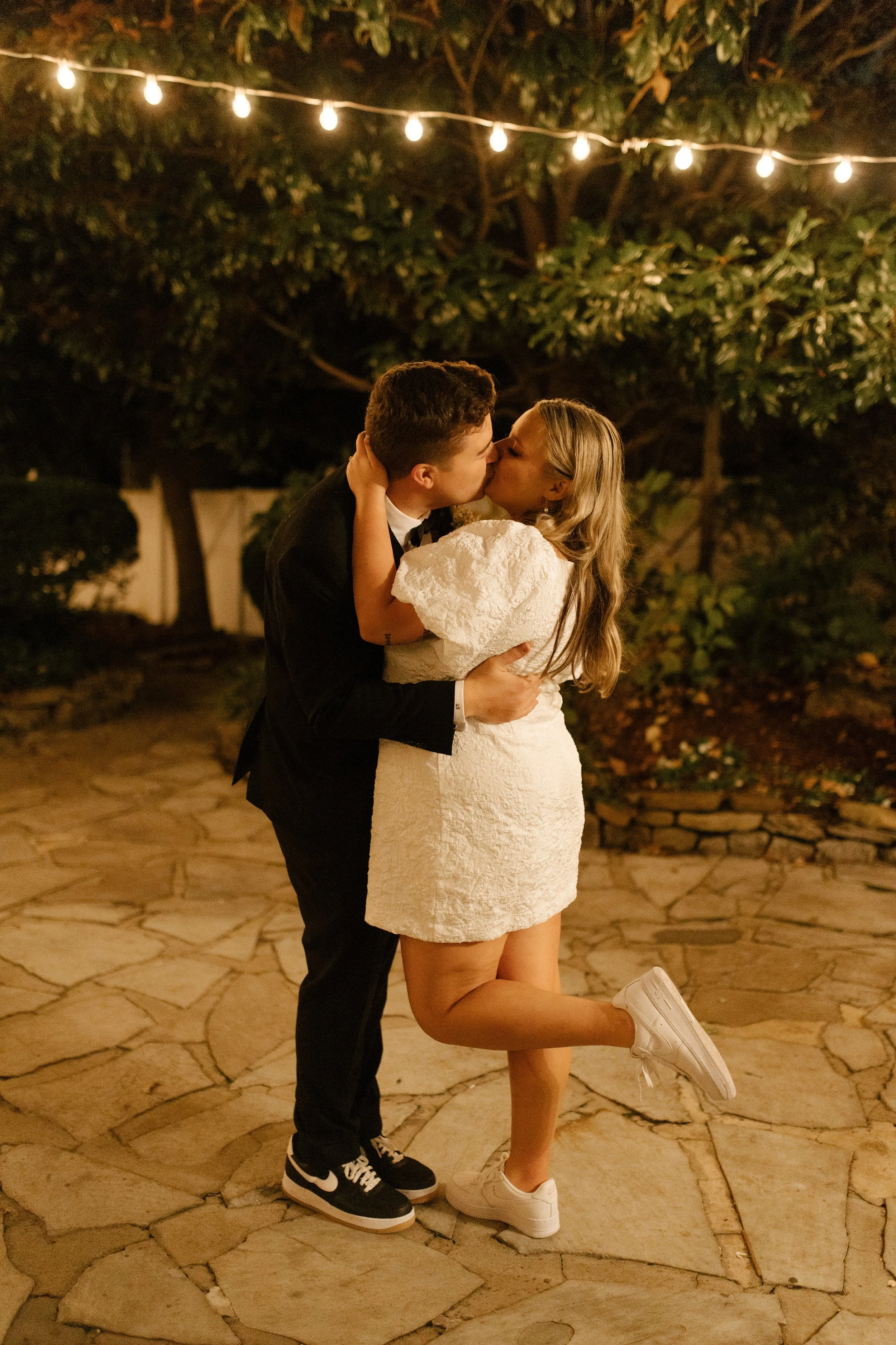 Newlyweds kissing beneath string lights in casual reception outfits and sneakers at the end of their wedding night.