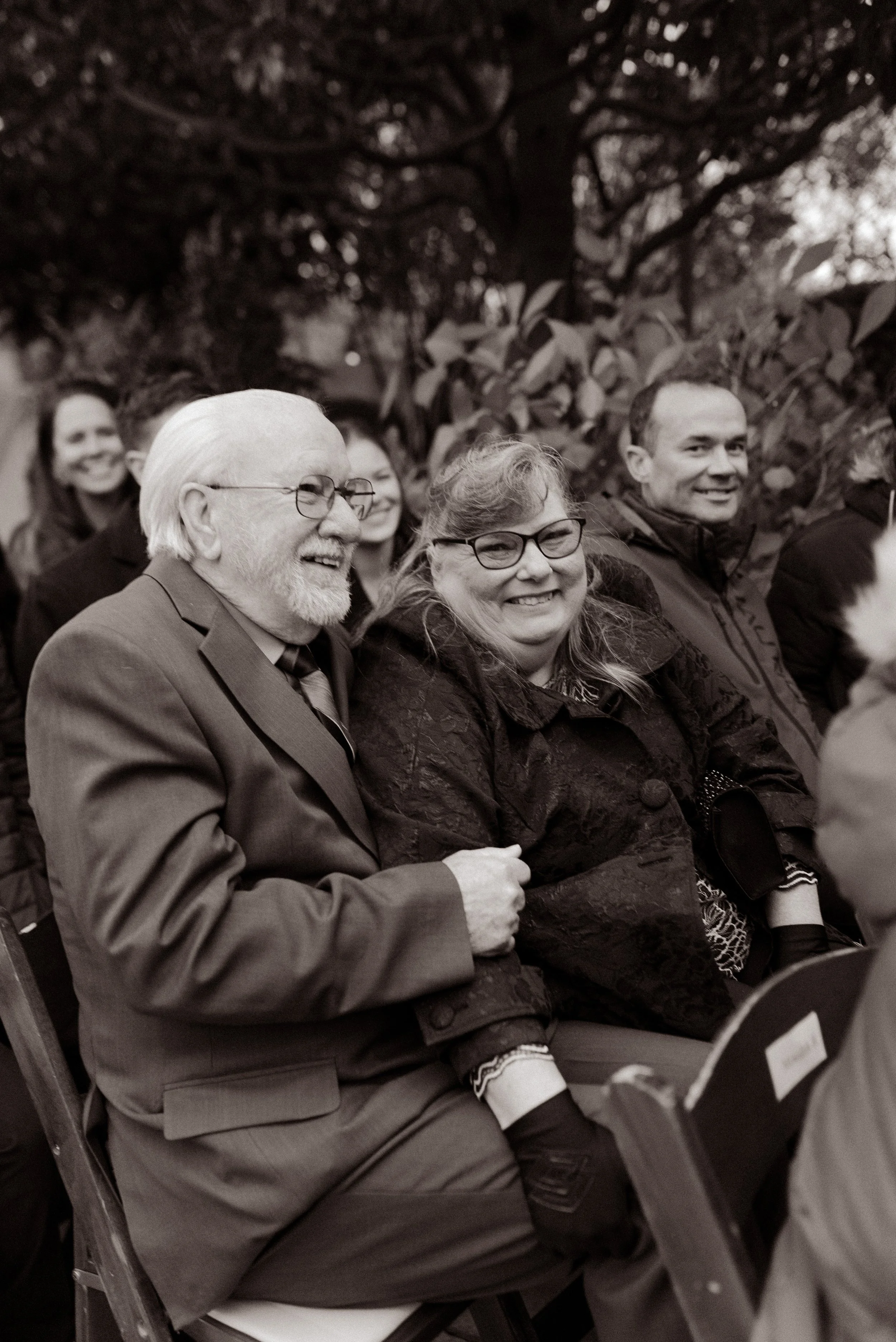 Black-and-white candid photo of smiling guests bundled up during an outdoor fall wedding ceremony.