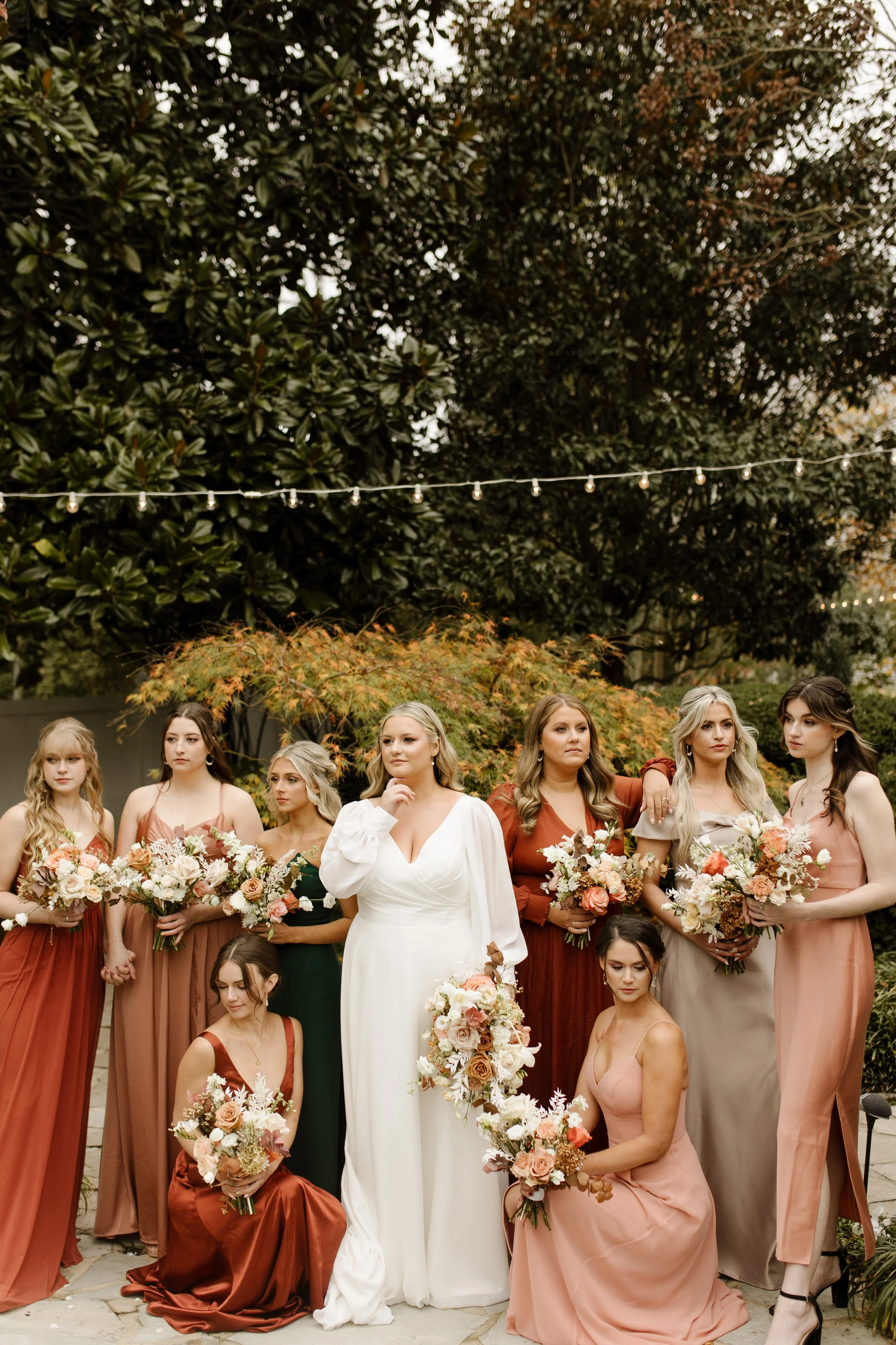 Bride surrounded by bridesmaids in rust, blush, green, and neutral dresses holding autumn-inspired bouquets at a fall wedding.